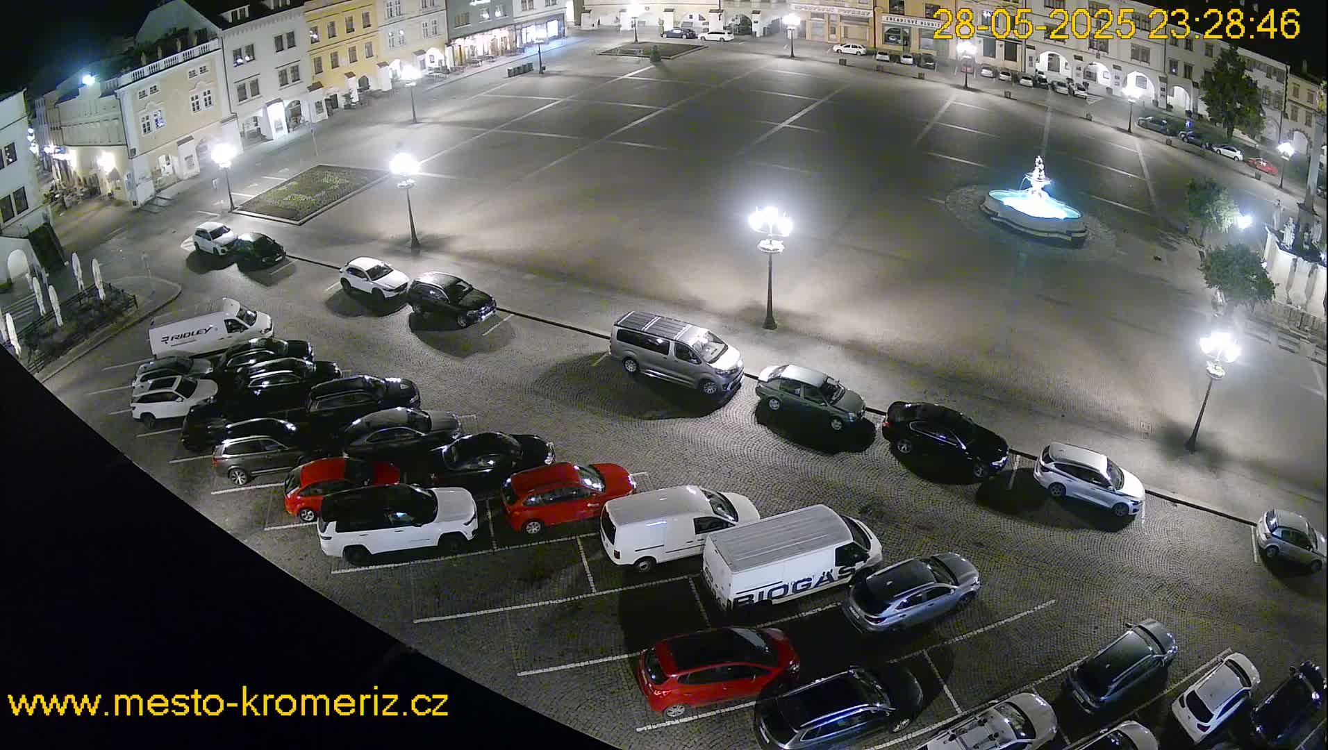 A nighttime, paved town square with a fountain at its center, surrounded by parked cars and buildings.
