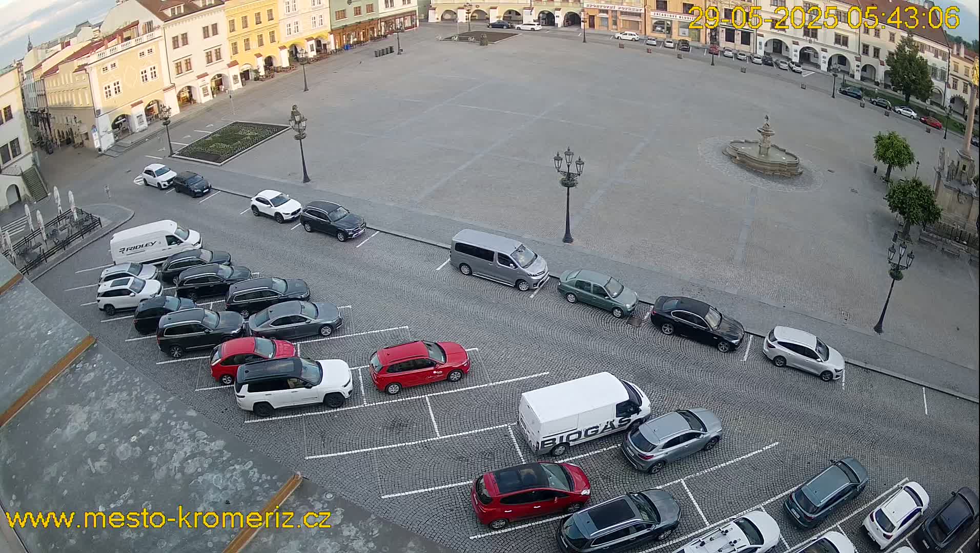 A town square with a fountain in the center is surrounded by buildings and numerous cars parked in spaces along the perimeter, under a partly cloudy sky.