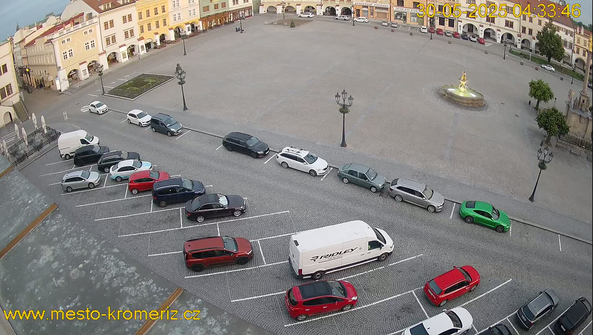 A town square with a fountain in the center, surrounded by buildings and parked cars, under a partly cloudy sky.