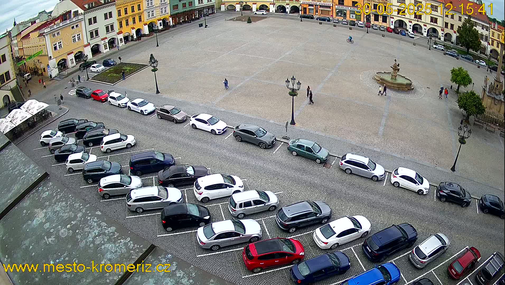An overcast day shows a town square with a fountain in the center and cars parked along its edges, surrounded by buildings.