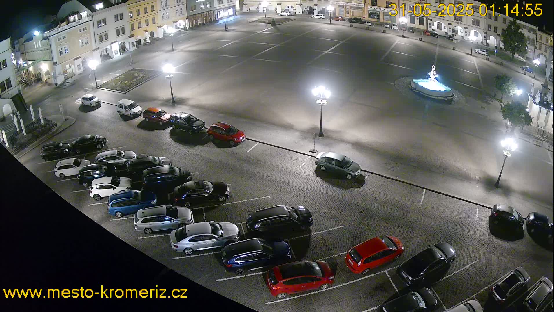 A nighttime, high-angle view of a town square with a fountain in the center, surrounded by parked cars and buildings.