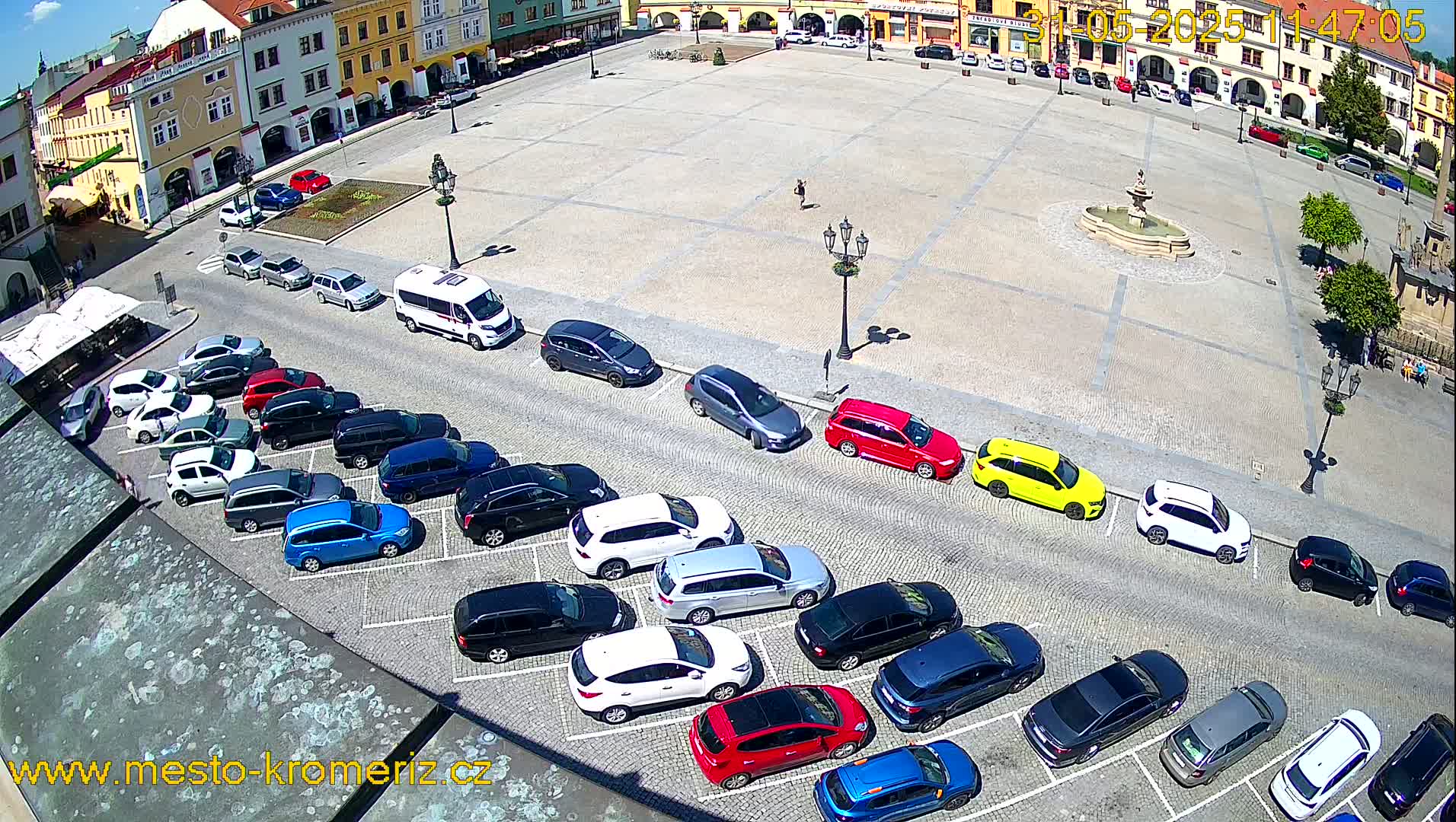 A large town square with a fountain in the center is surrounded by buildings, and numerous cars are parked along the sides of the square on a sunny day.