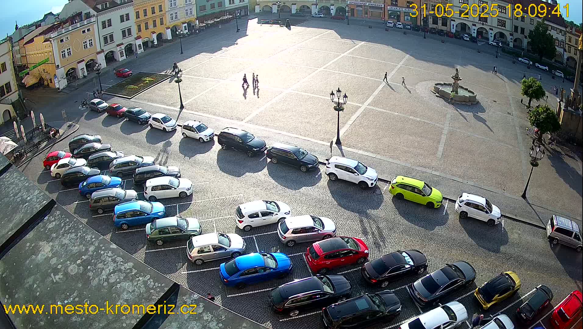 A sunny day in a town square shows a cobblestone area with a fountain, several pedestrians, and numerous cars parked along the edges.