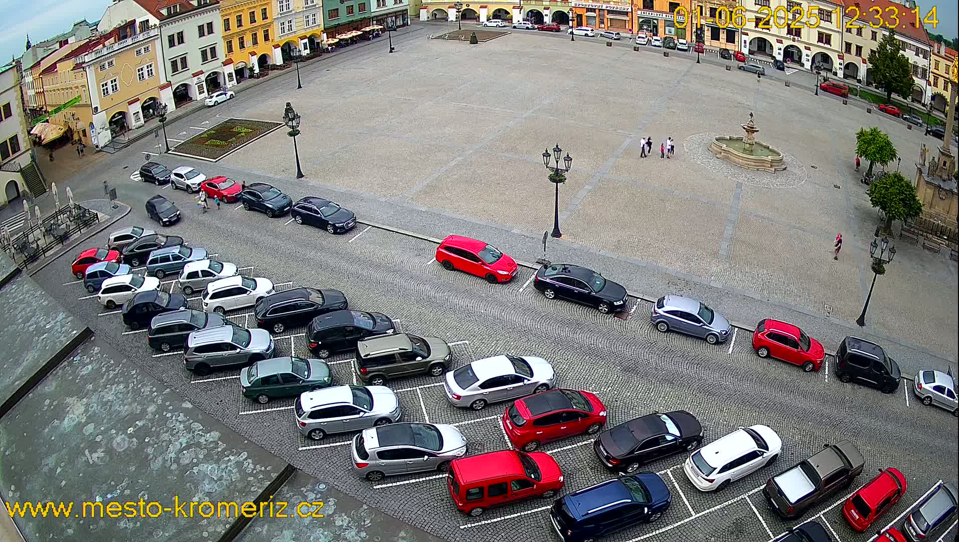 A large town square with a fountain in the center, surrounded by parked cars and buildings, on an overcast day.