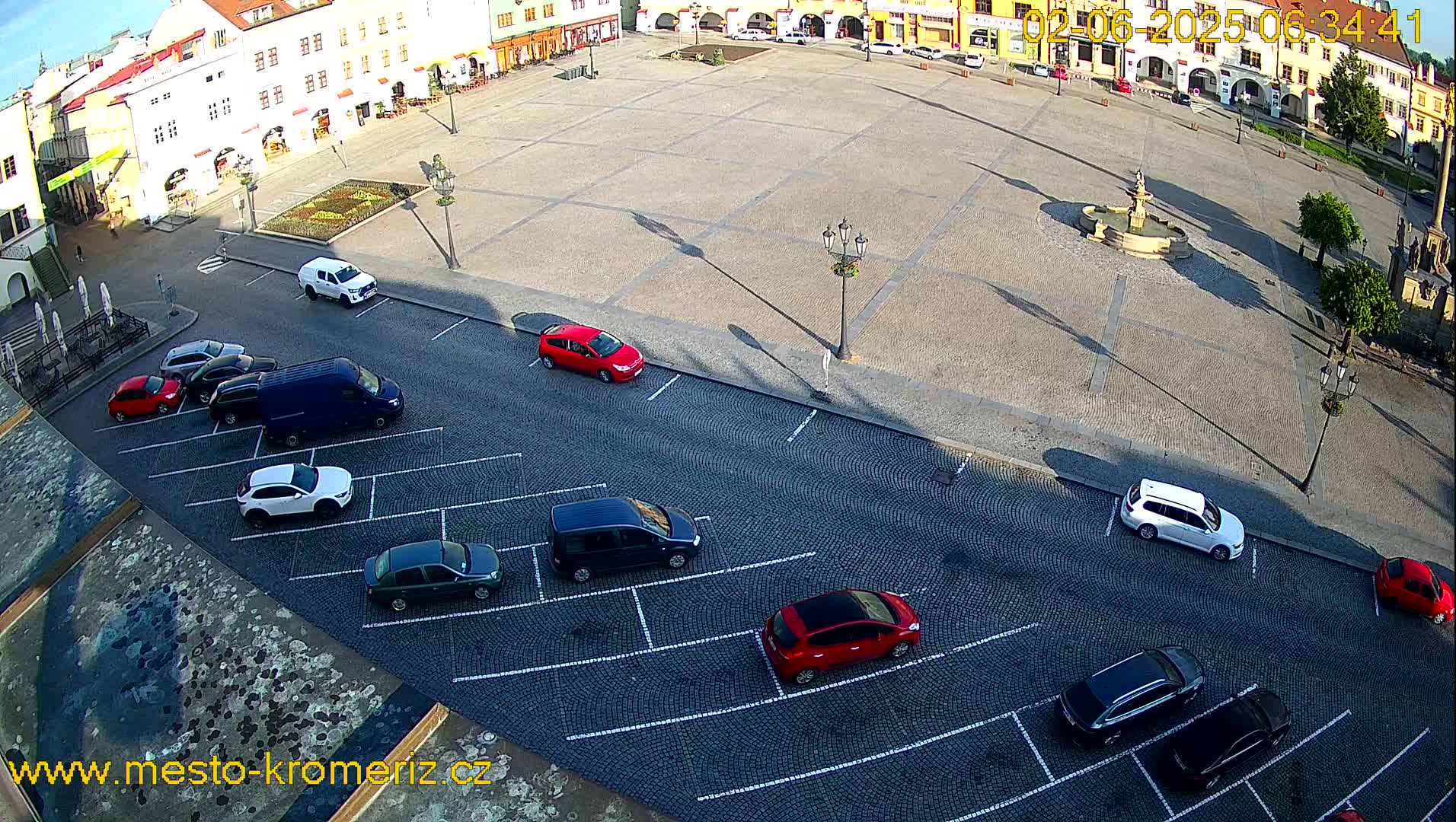 A paved town square with a fountain in the center is surrounded by buildings, and several cars are parked along the street adjacent to the square on a sunny day.