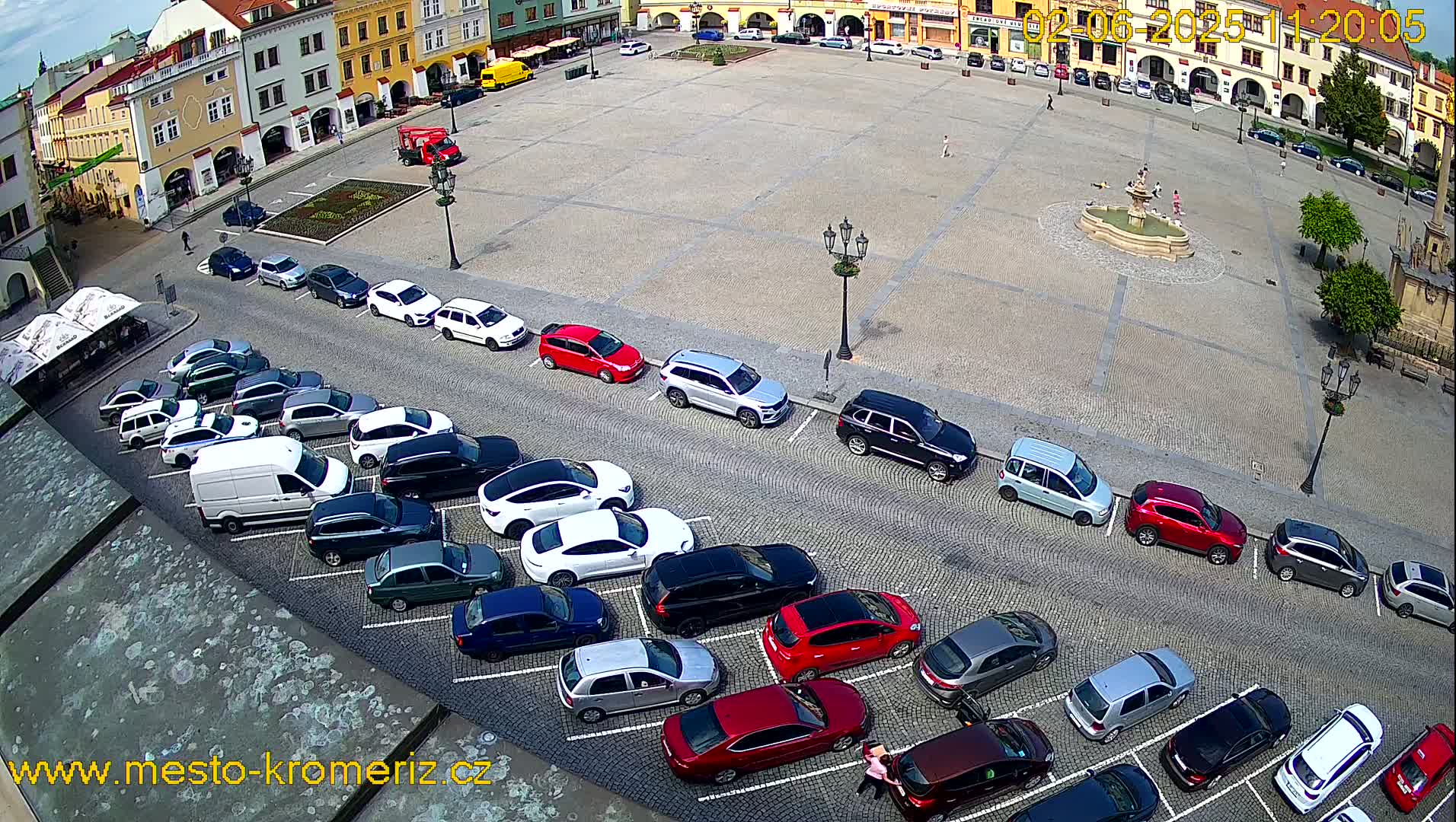 A large town square with a fountain in the center is surrounded by buildings and has cars parked along its edges on a sunny day.