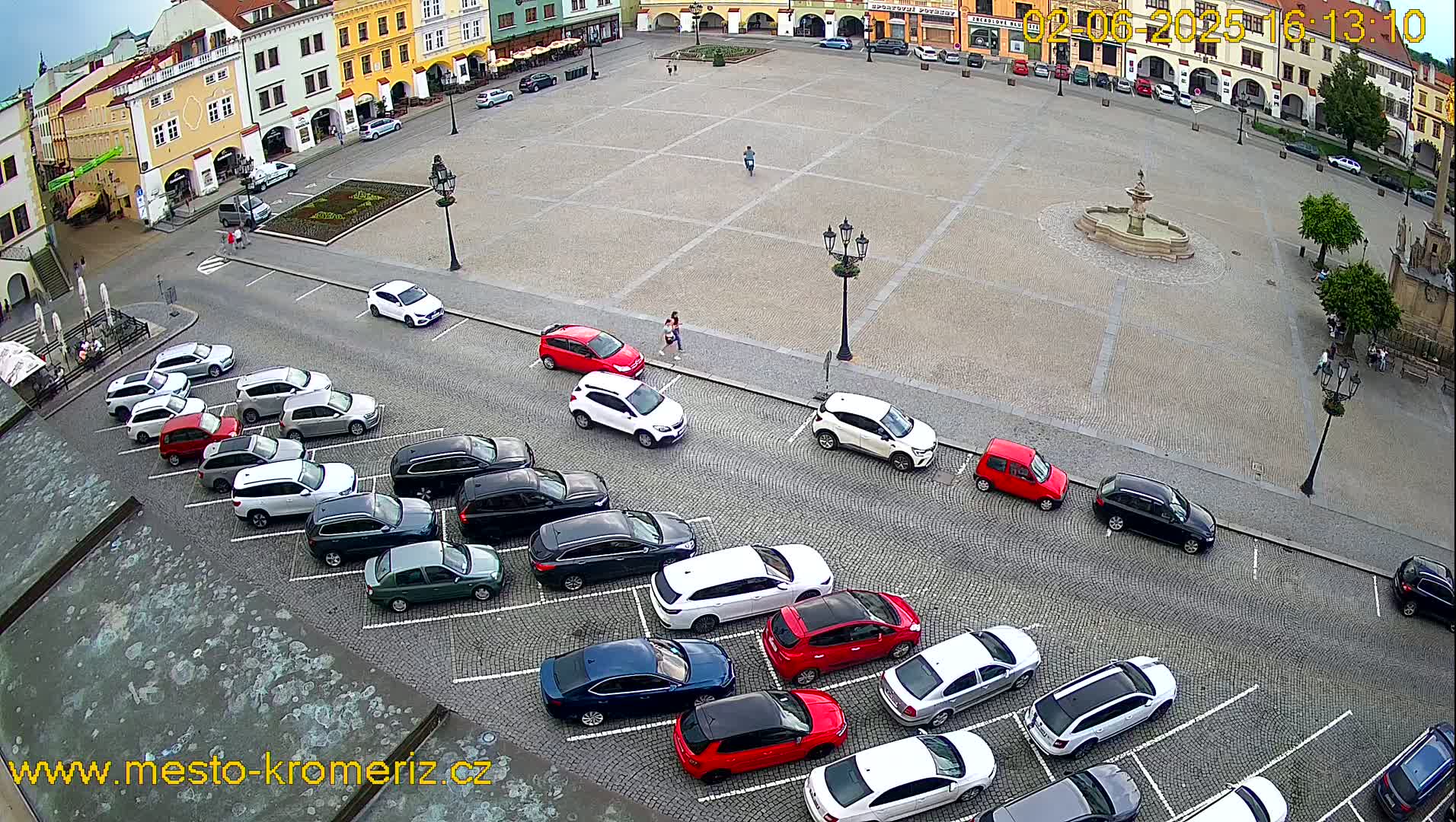 An aerial view shows a town square with a fountain in the center, surrounded by buildings and numerous cars parked along the edges on a partly sunny day.