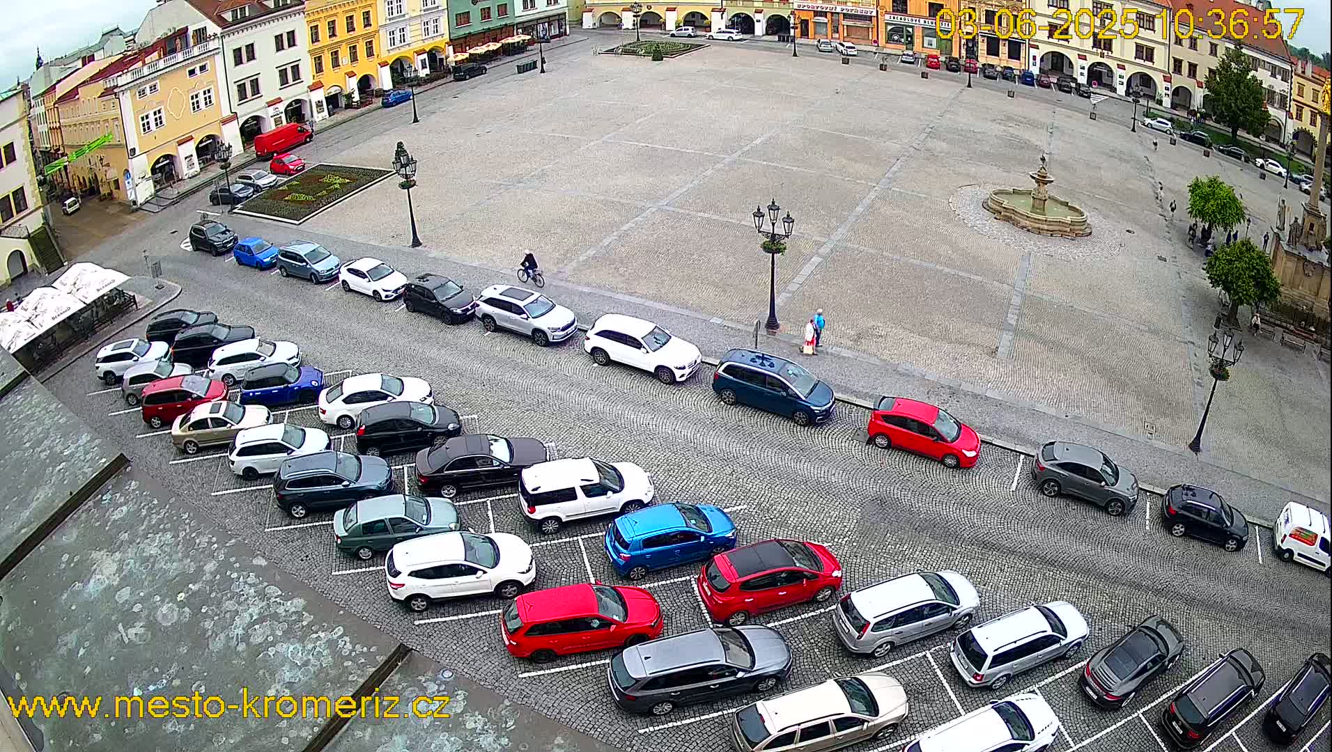 A large town square with a fountain in the center is surrounded by buildings, and numerous cars are parked along the sides.