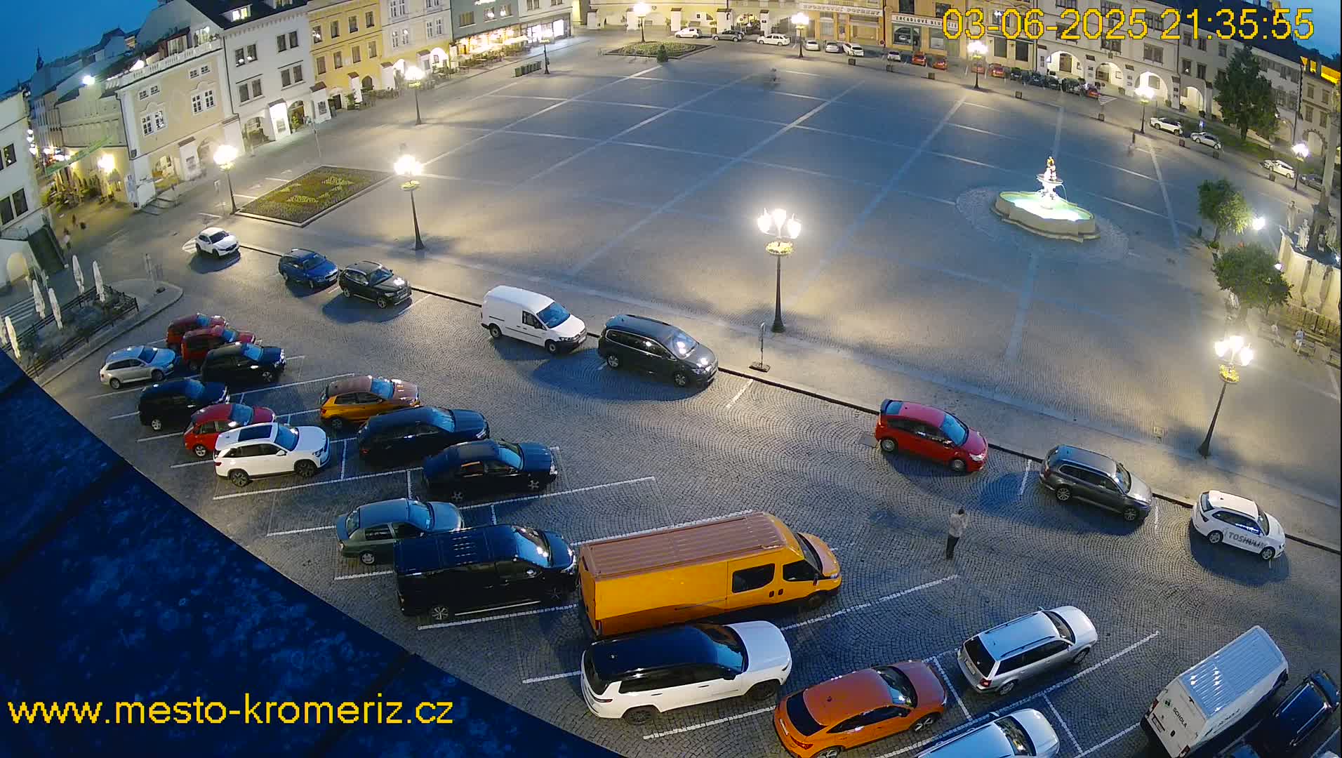 A large, empty square at night with cars parked along the edges, lit by streetlights, and a fountain in the center.
