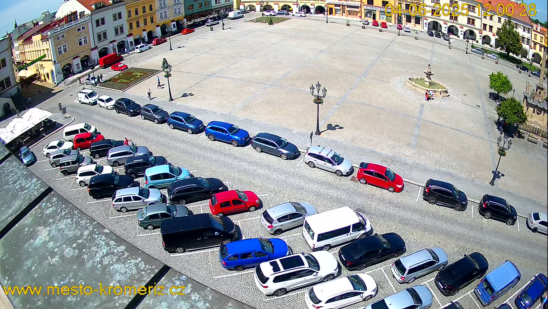 A town square with a fountain in the center is surrounded by parked cars and buildings on a sunny day.