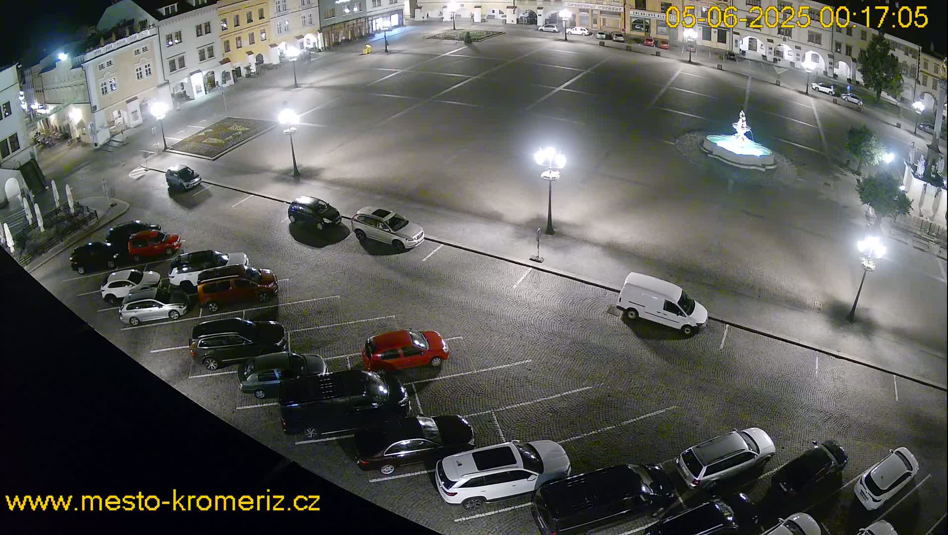 A nighttime aerial view of a town square with a fountain at its center, surrounded by parked cars and buildings illuminated by streetlights.