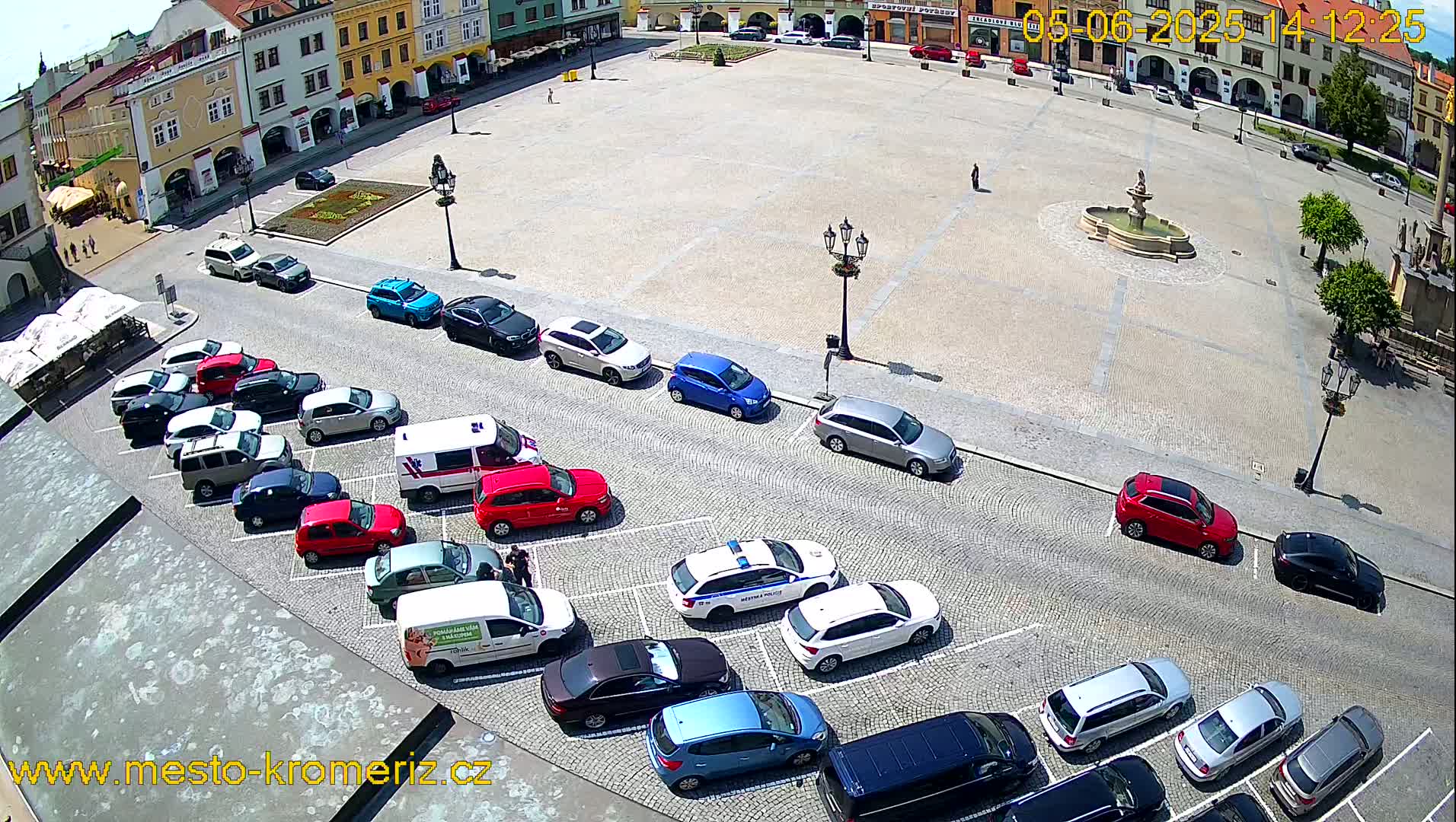 A sunny day in a town square with a fountain, surrounded by buildings, and numerous cars parked along the edges.