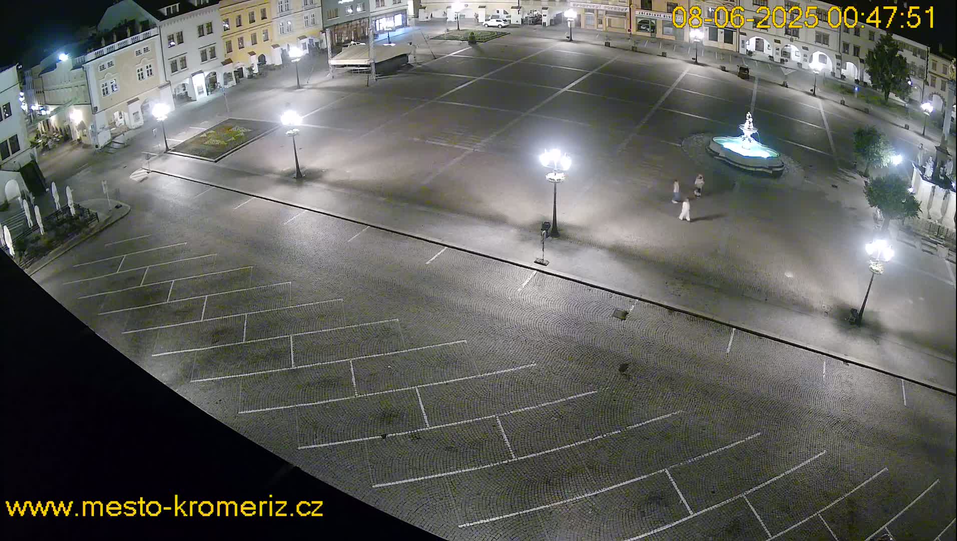A nighttime, aerial view of a cobblestone town square with a fountain, parking spaces, and several lampposts illuminating the mostly empty space.
