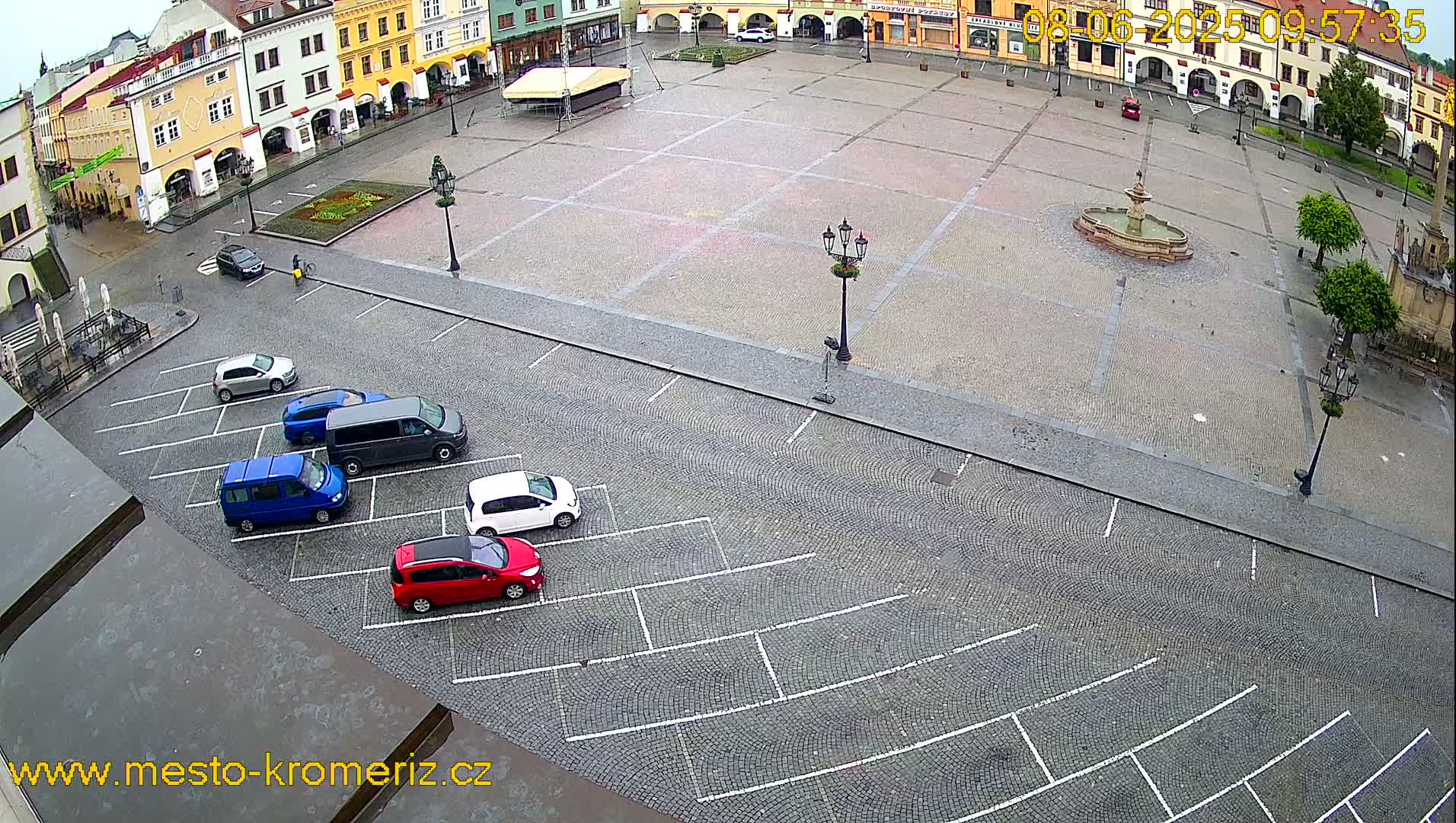 A cobblestone town square with a fountain, several parked cars, and surrounding buildings is visible on an overcast day.