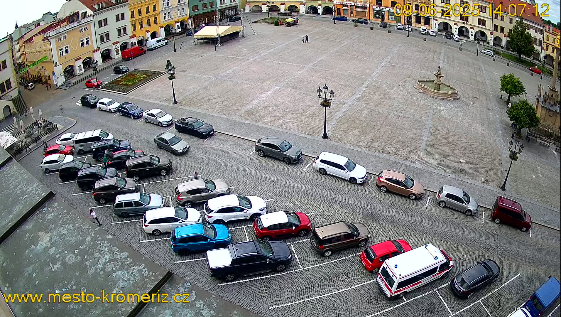 A town square with a cobblestone surface, a central fountain, and cars parked along the edges, under overcast skies.