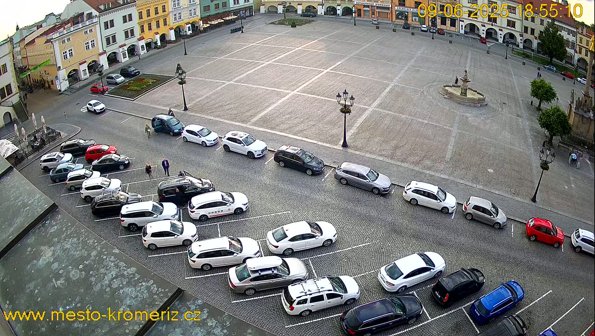 A town square with a fountain in the center, surrounded by buildings, and numerous cars parked along the sides under a partly cloudy sky.