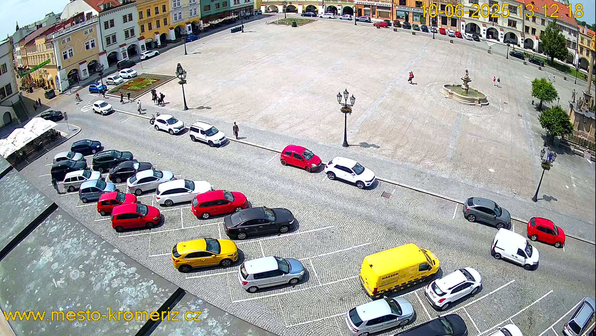 A large town square with a fountain and many parked cars is viewed from above on a sunny day.