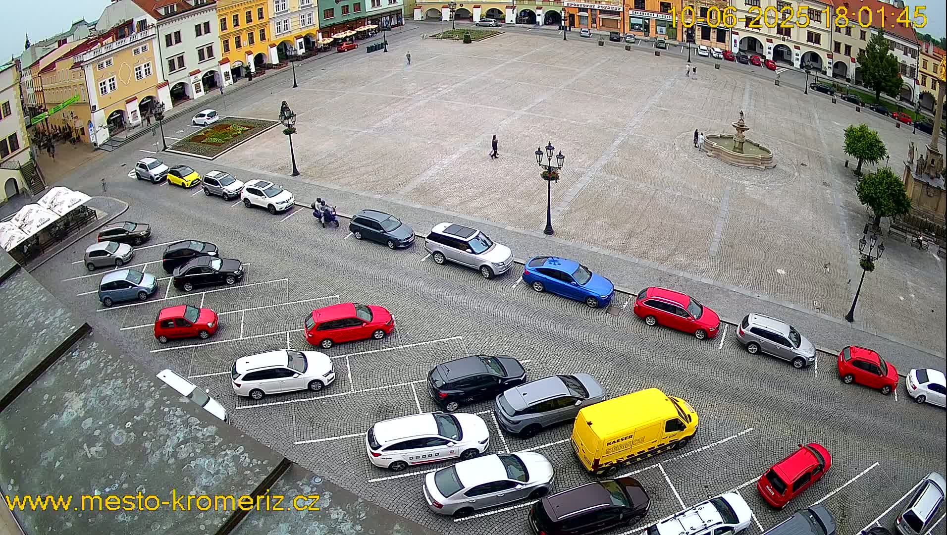 A large town square with a fountain at its center is surrounded by buildings and parked cars, under an overcast sky.