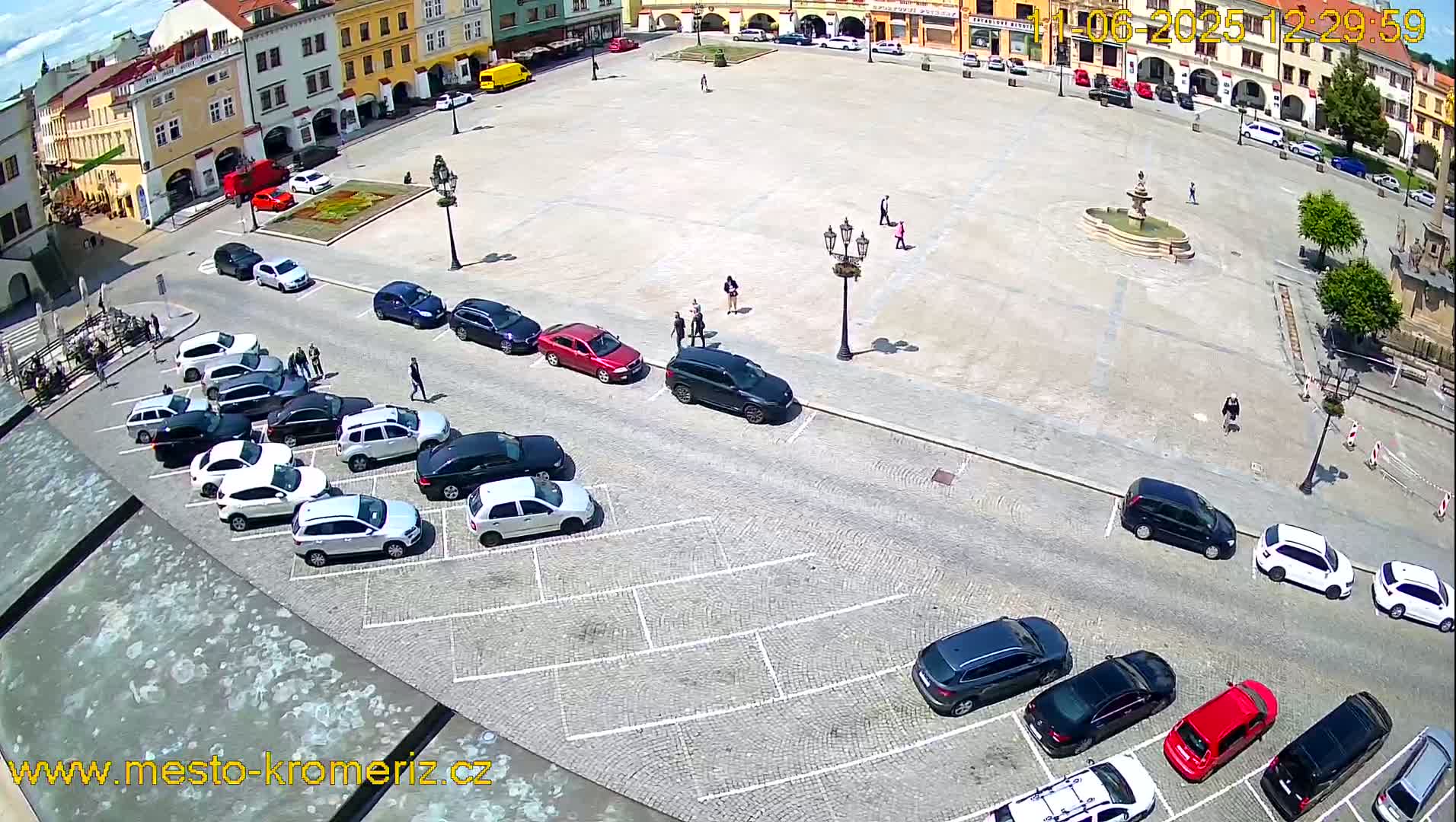 A large town square, under sunny skies, is surrounded by buildings and has several cars parked along its edge.