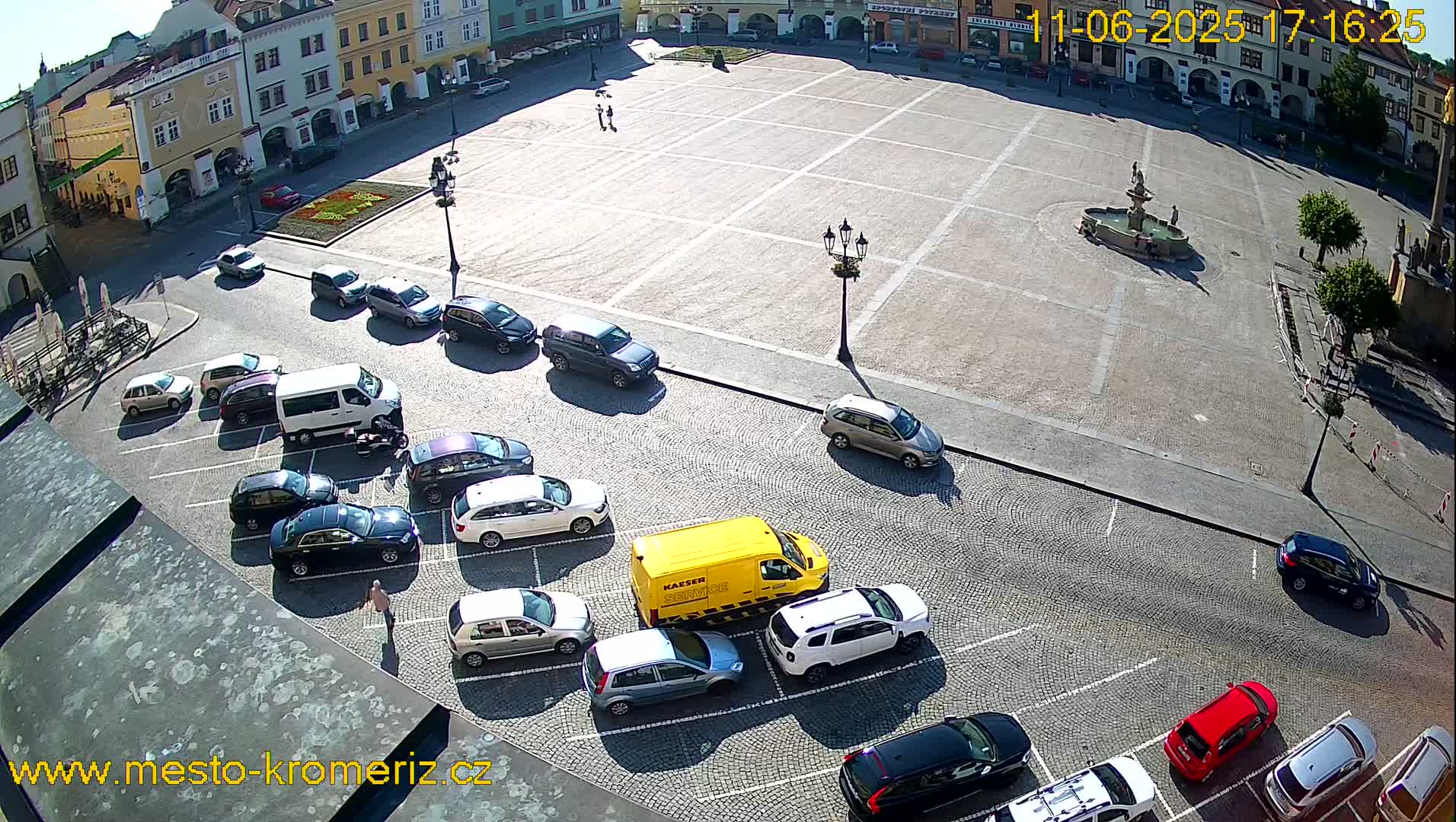 A large, paved town square with a fountain in the center is surrounded by parked cars and buildings under sunny conditions.