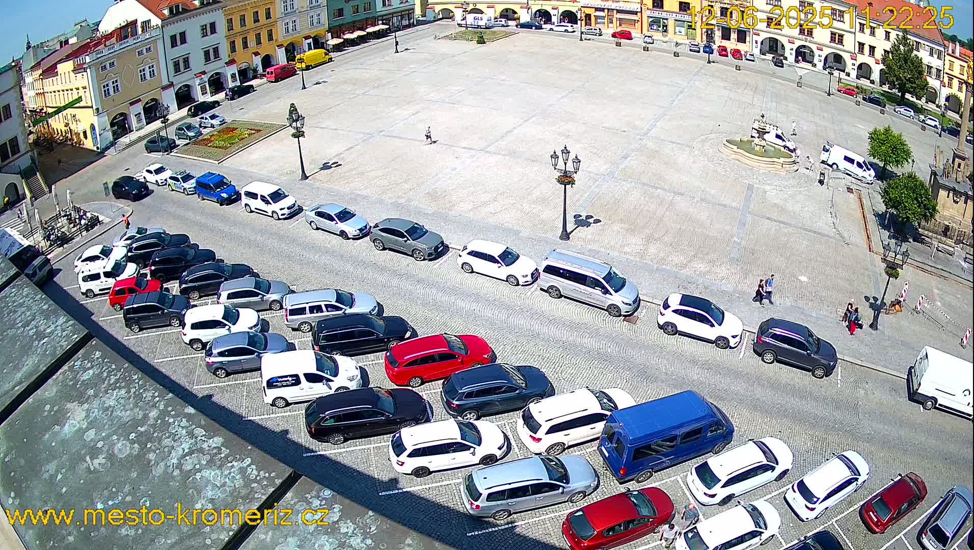 A sunny day shows a large town square with many cars parked along its sides, a fountain in the center, and several people walking around.