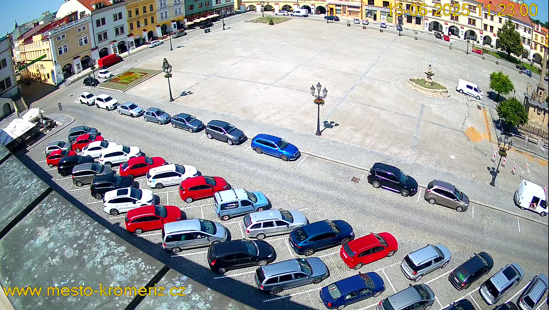 A sunny day reveals a large town square with numerous cars parked in parallel rows along its edges, surrounding a central fountain.