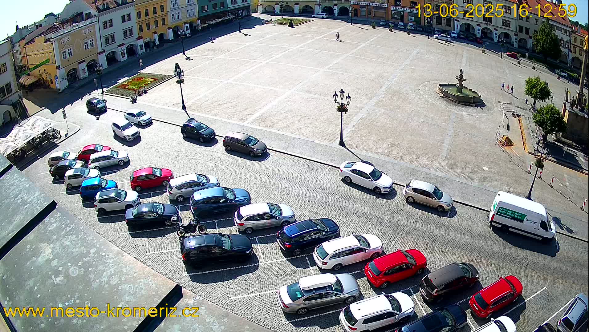 A high-angle view shows a town square on a sunny day with cars parked along one side and a fountain in the center.