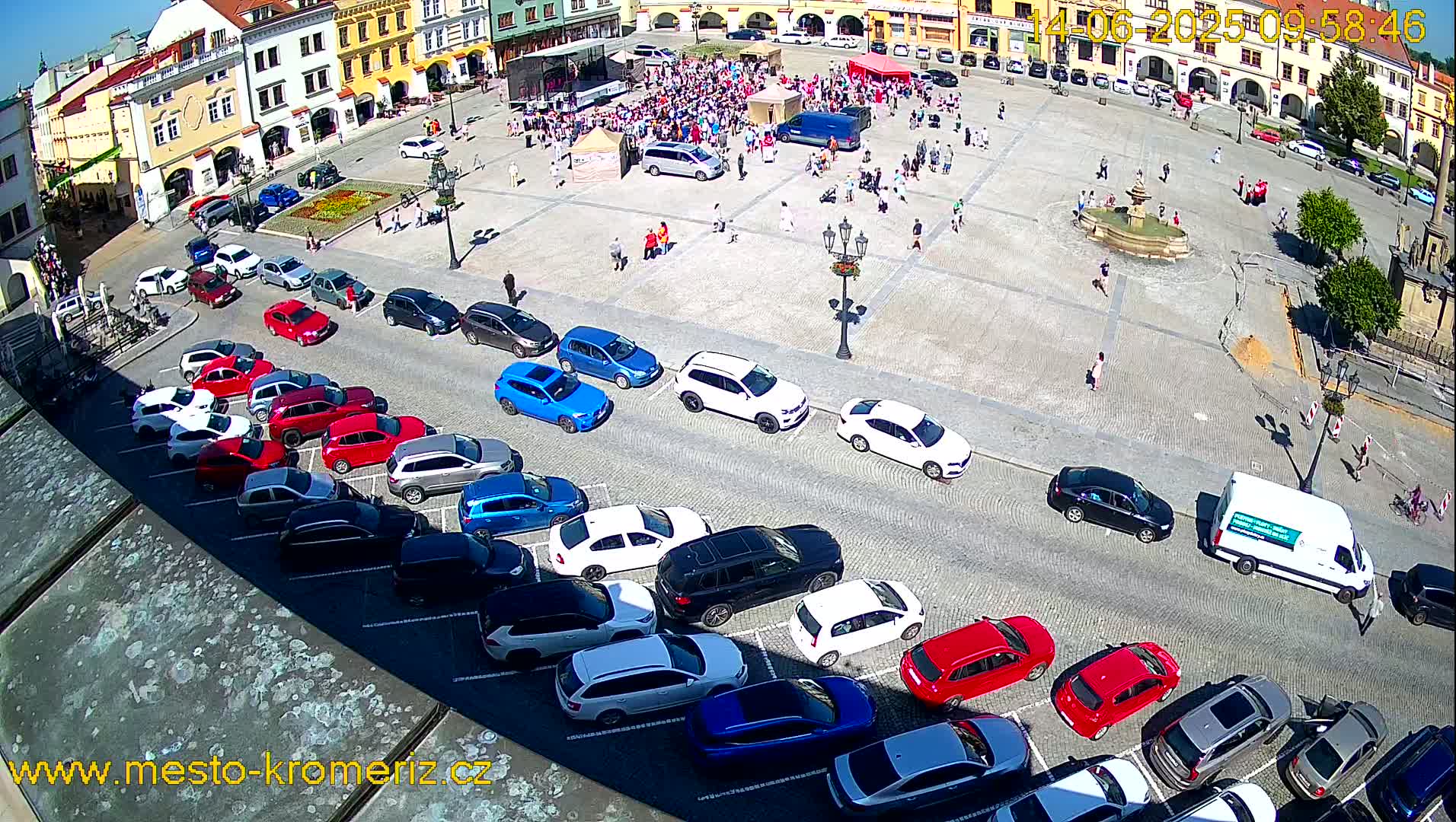 A town square on a sunny day, filled with parked cars and people gathered in the center.