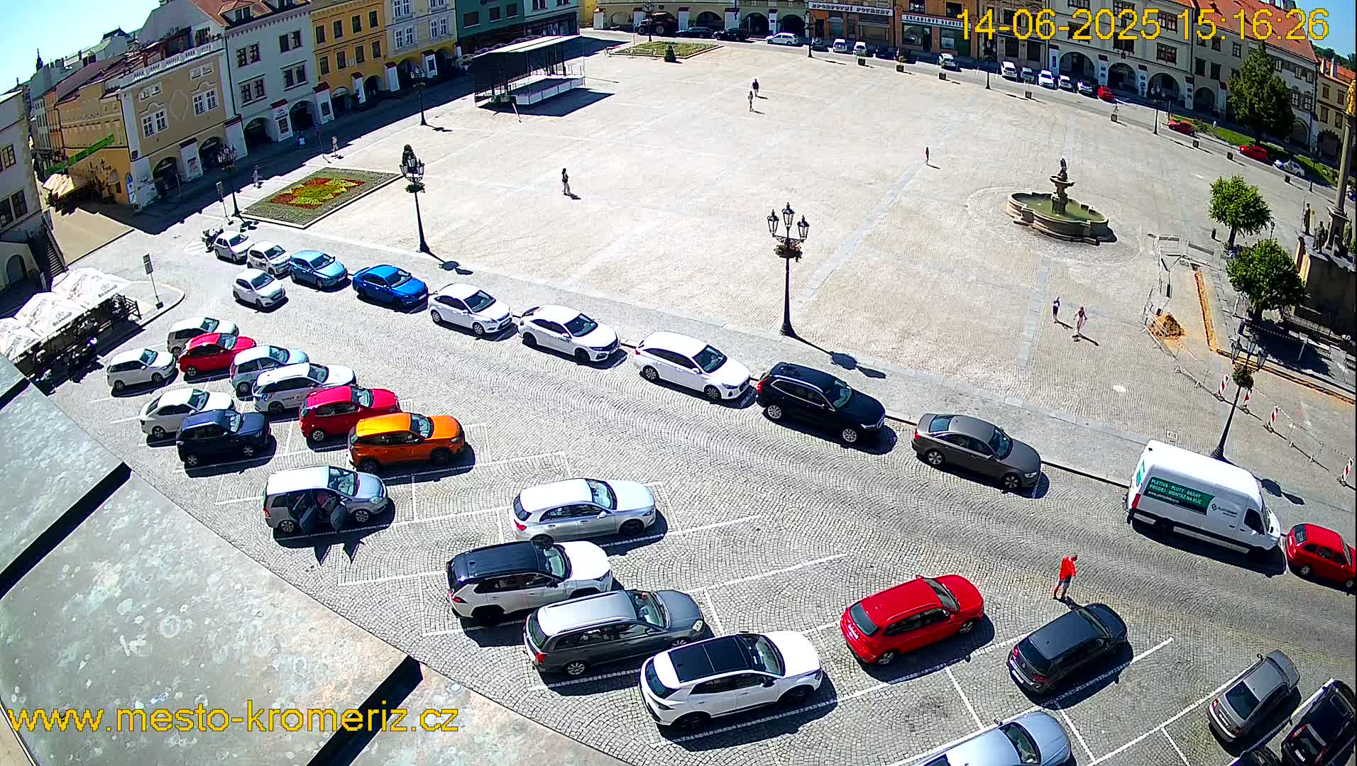 A sunny day shows a town square with a fountain in the center and multiple cars parked in rows along the edges.