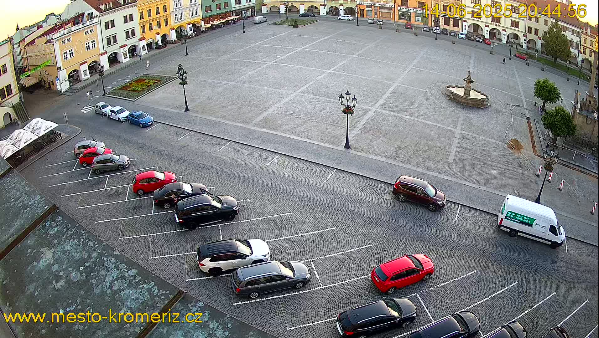 A sunny day shows a town square with a fountain in the center and multiple cars parked in rows along the edges.