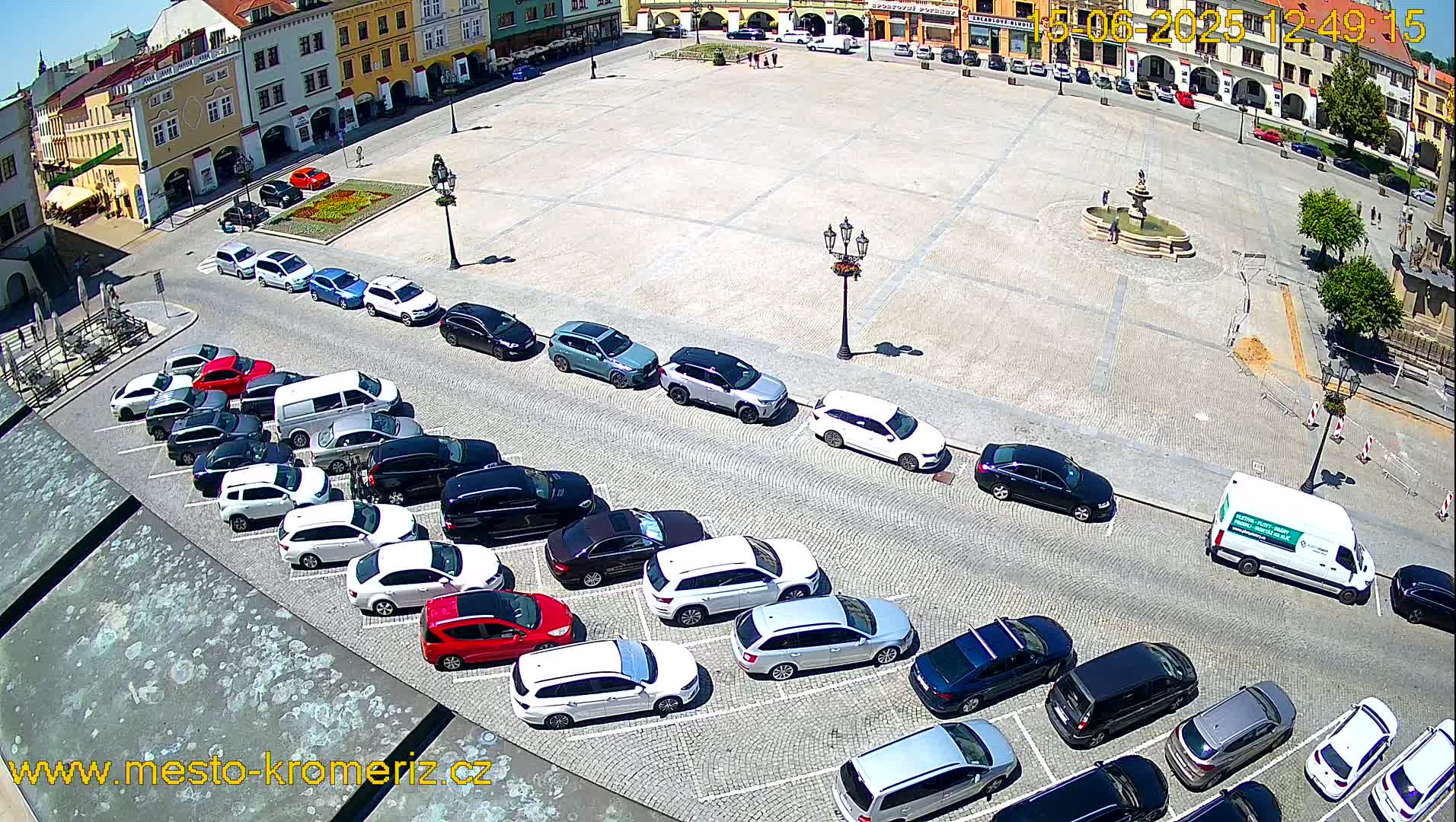 A large town square on a sunny day is filled with cars parked along its edges, and a fountain sits in the center.