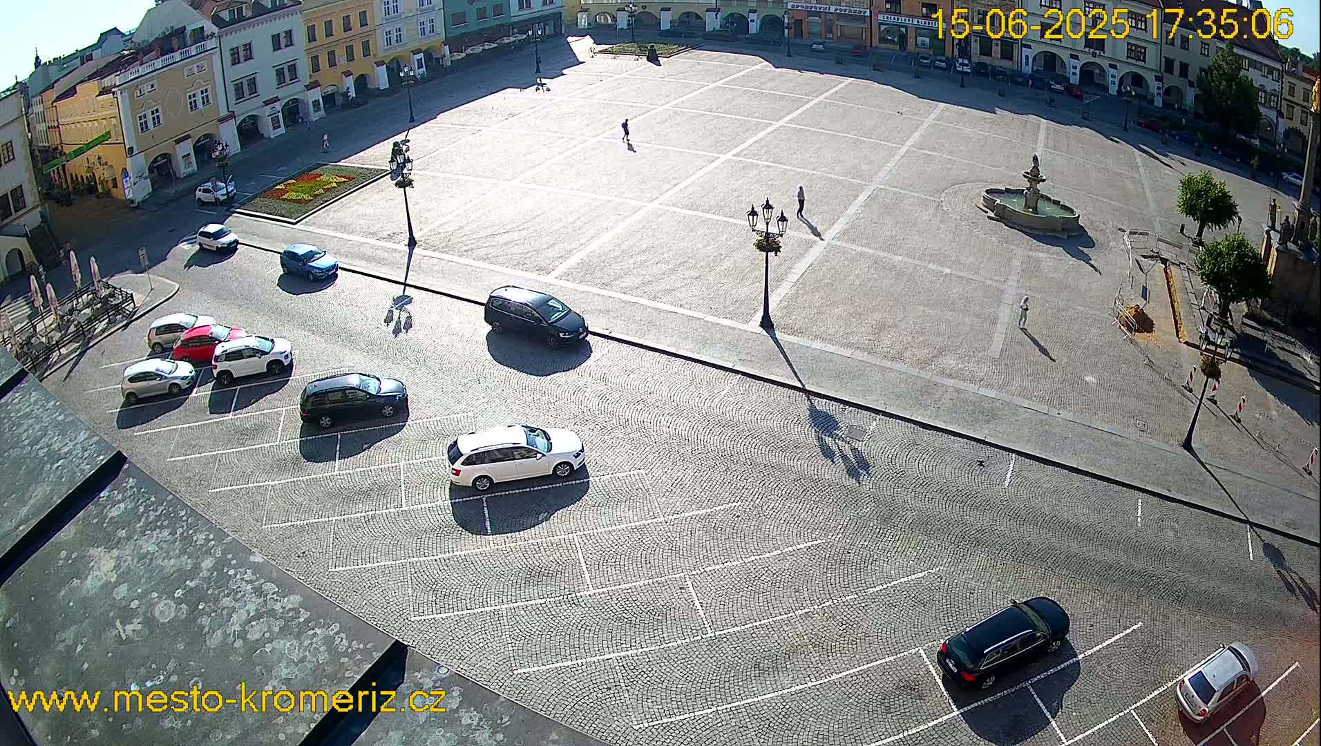 A sunny day reveals a large cobblestone town square with a fountain, several parked cars, and a few pedestrians.