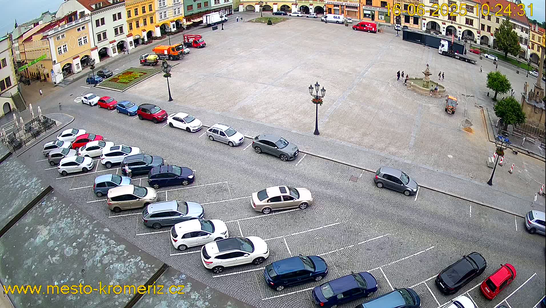 A paved town square with a fountain and numerous cars parked along the sides, under an overcast sky.