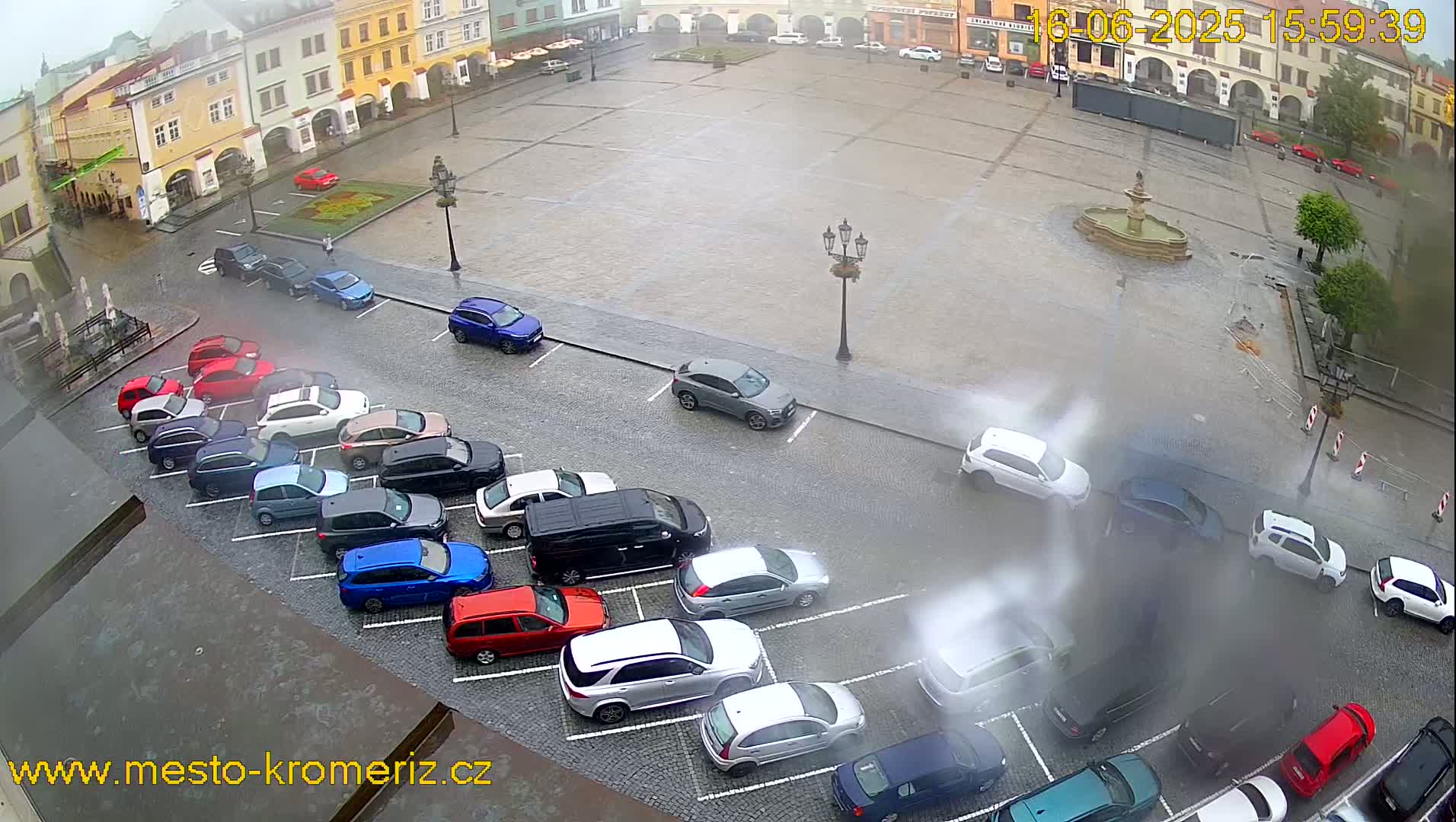 A rain-soaked town square with parked cars lining the edges and a fountain in the center.
