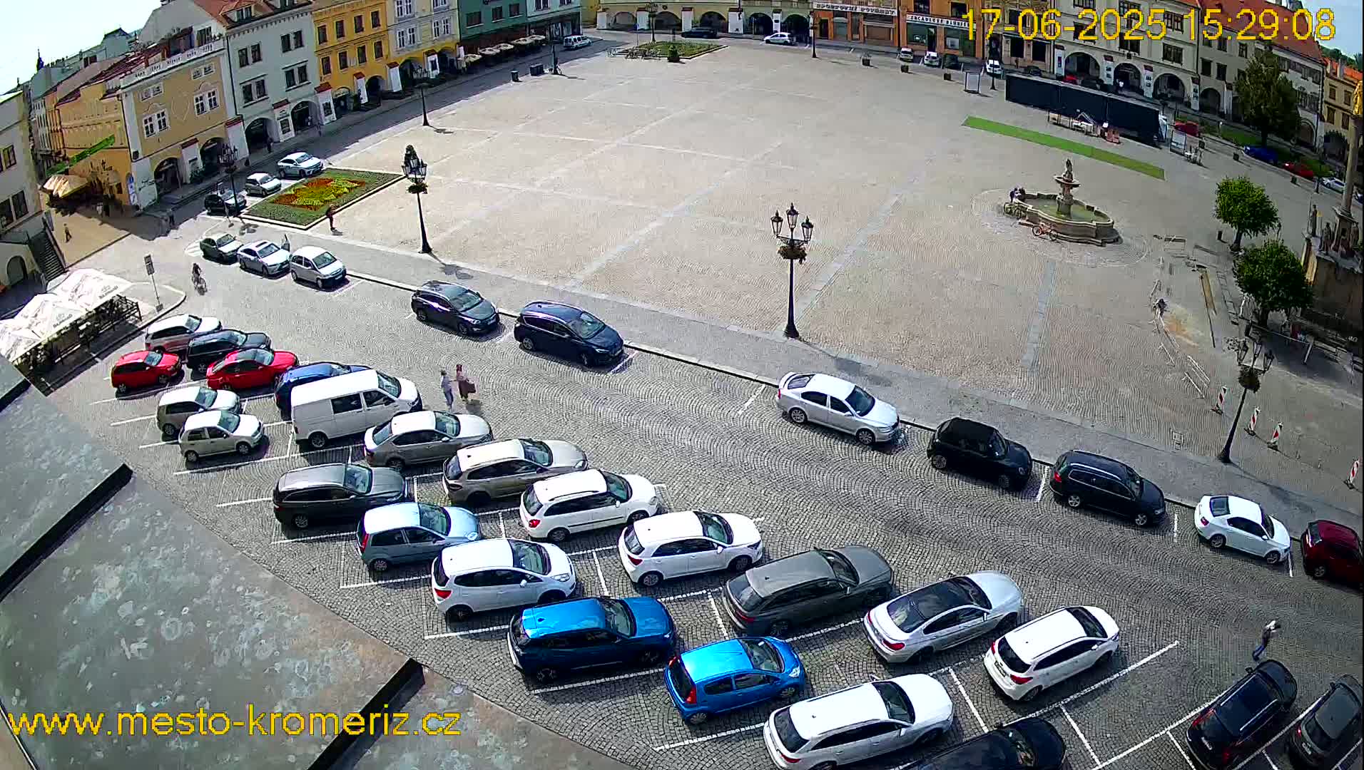 A town square with a fountain in the center, surrounded by buildings and cars parked along the sides on a sunny day.