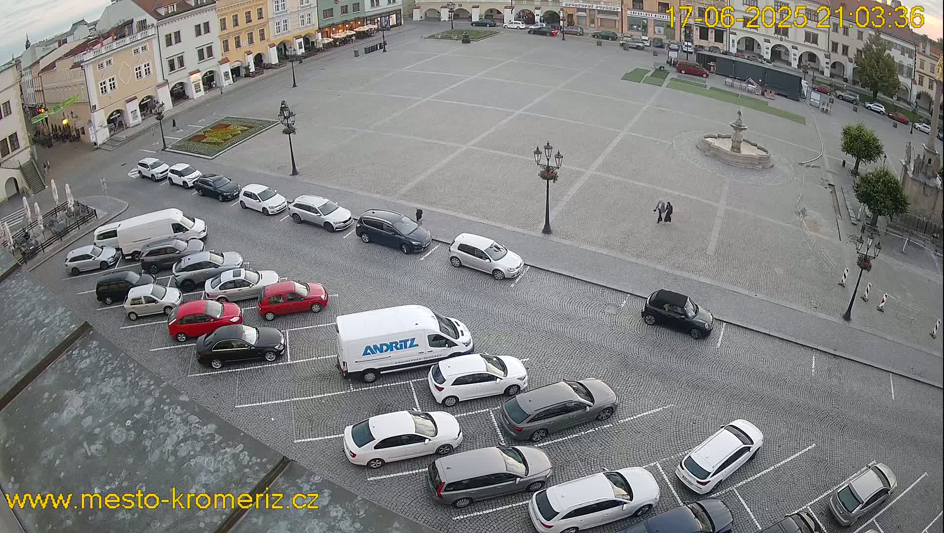 A large town square, mostly empty except for parked cars and a few pedestrians, is surrounded by buildings under a partly cloudy evening sky.