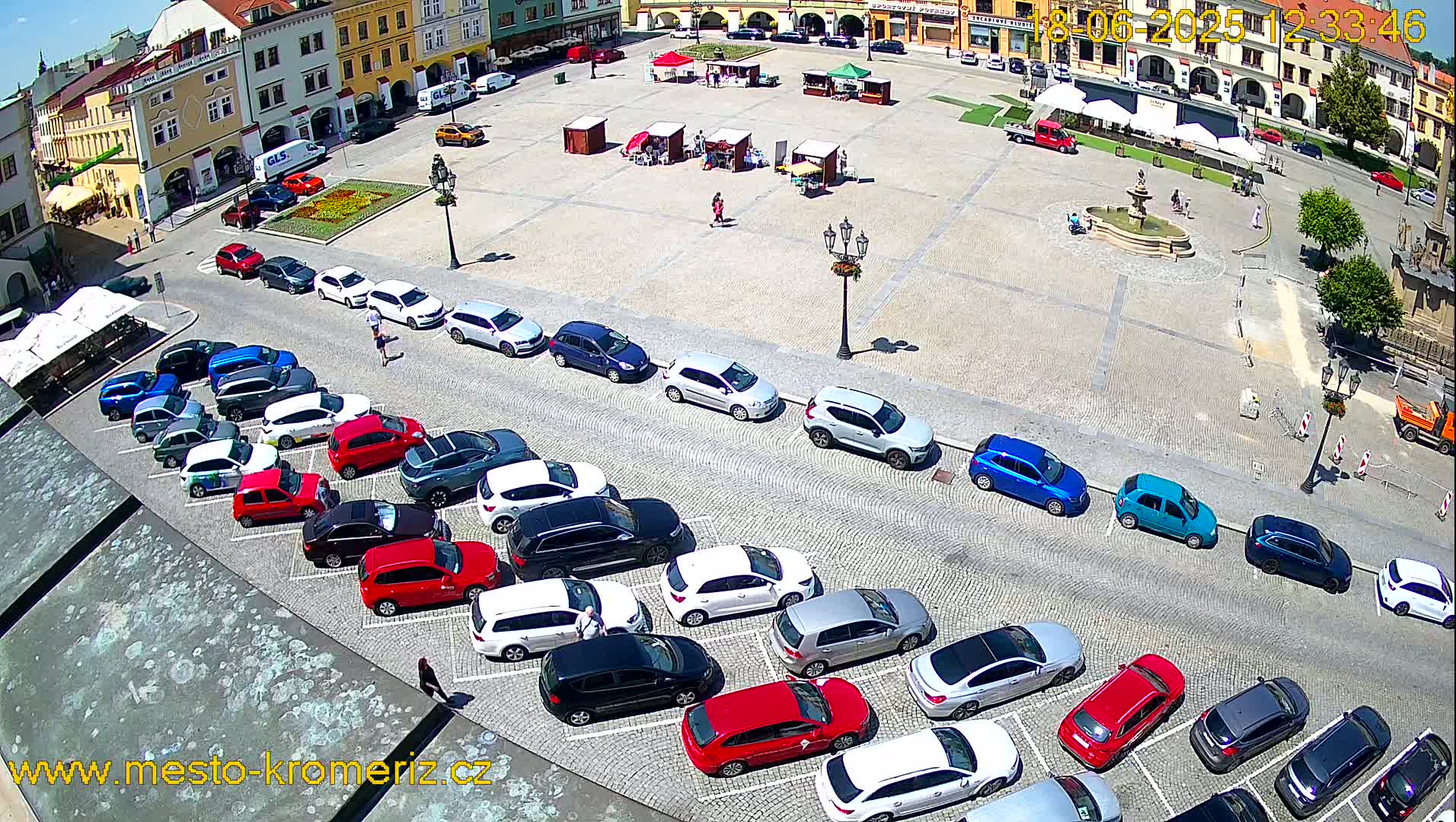 A sunny day shows a town square with several cars parked in rows along its perimeter, and a small fountain in the center.