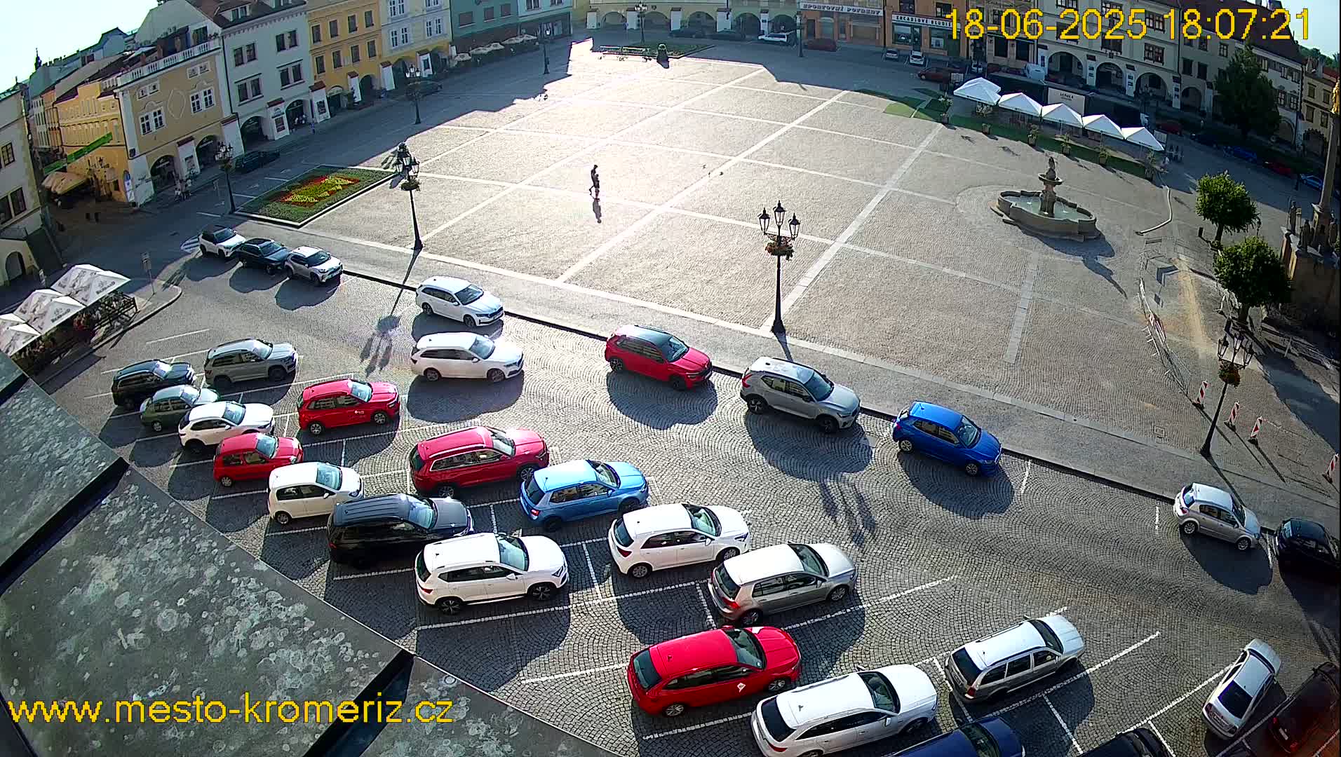 A sunny day reveals a cobblestone town square with several cars parked along its edges, a fountain at its center, and buildings surrounding it.