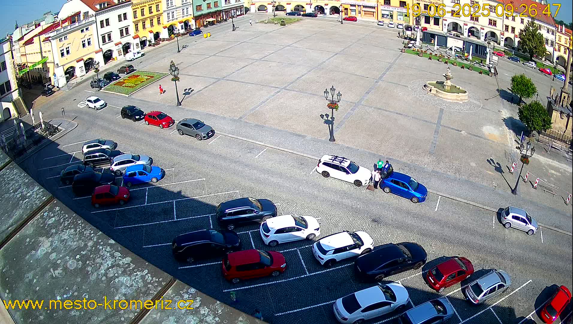 A sunny day shows a large town square with a fountain in the center, surrounded by buildings and cars parked along the sides.