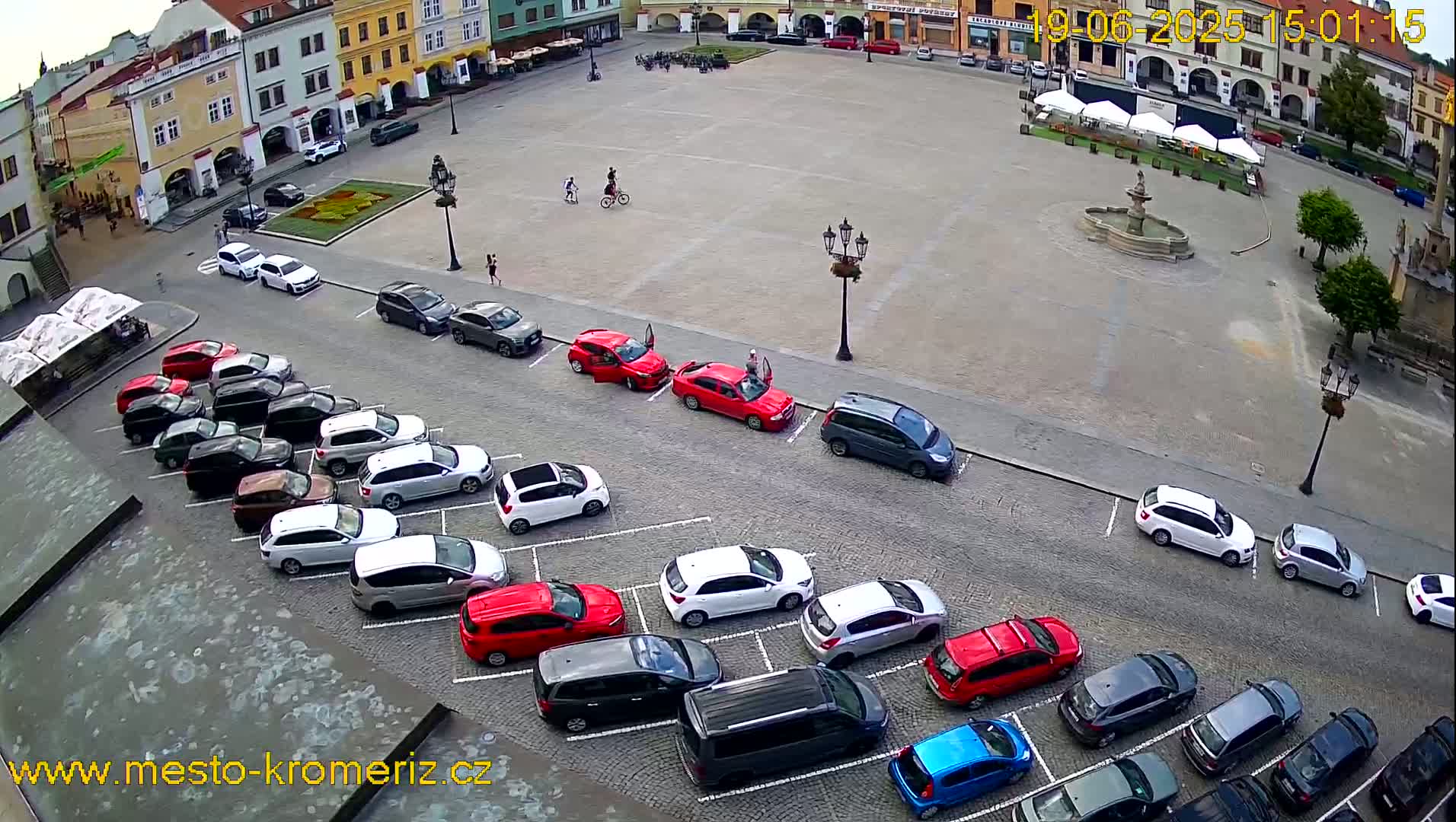 A town square with a fountain, surrounded by buildings and numerous cars parked in a lot adjacent to the square, on a mostly sunny day.