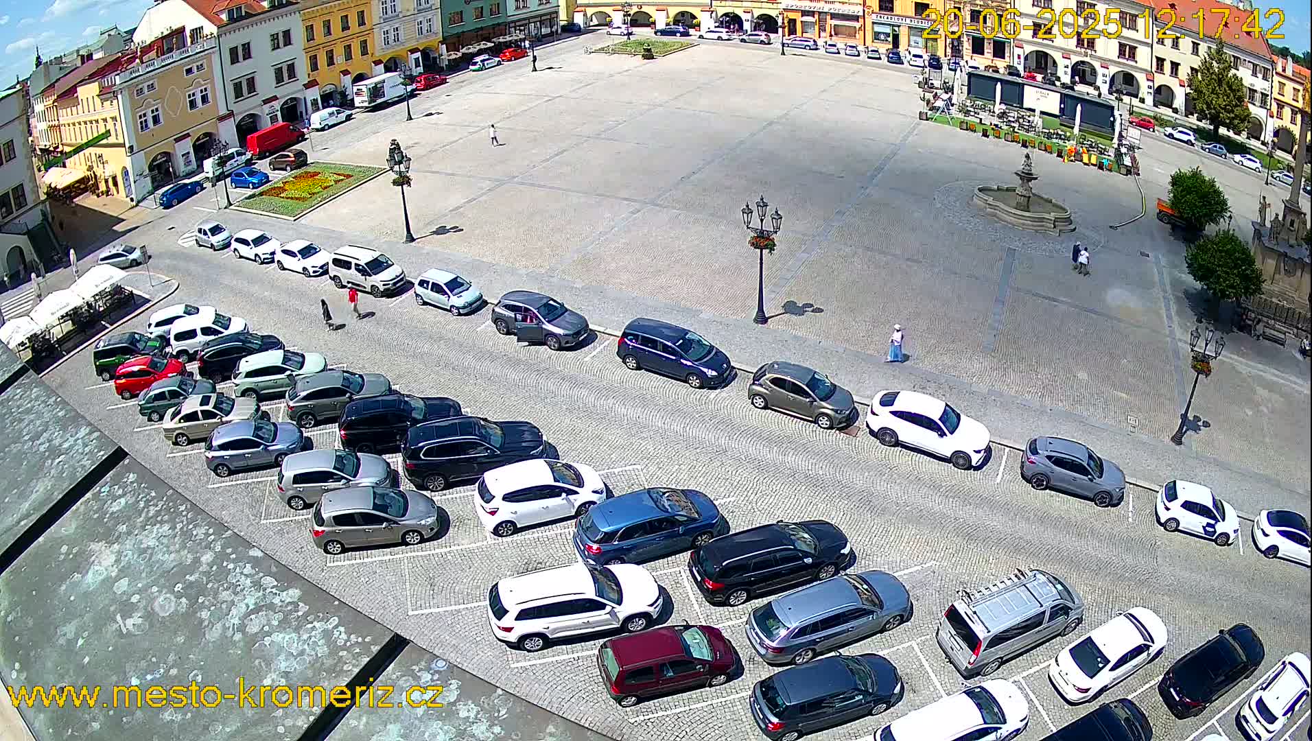 A large town square with a fountain in the center, surrounded by parked cars and buildings, on a sunny day.