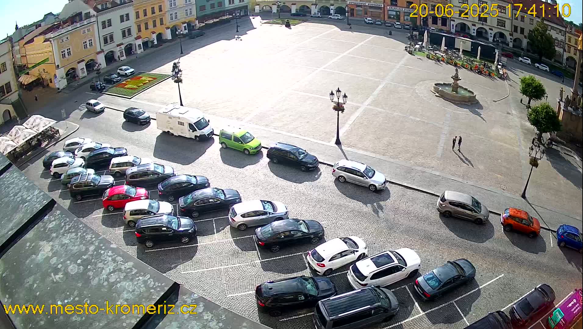 A sunny day shows a town square with a fountain and many cars parked along the sides, viewed from above.