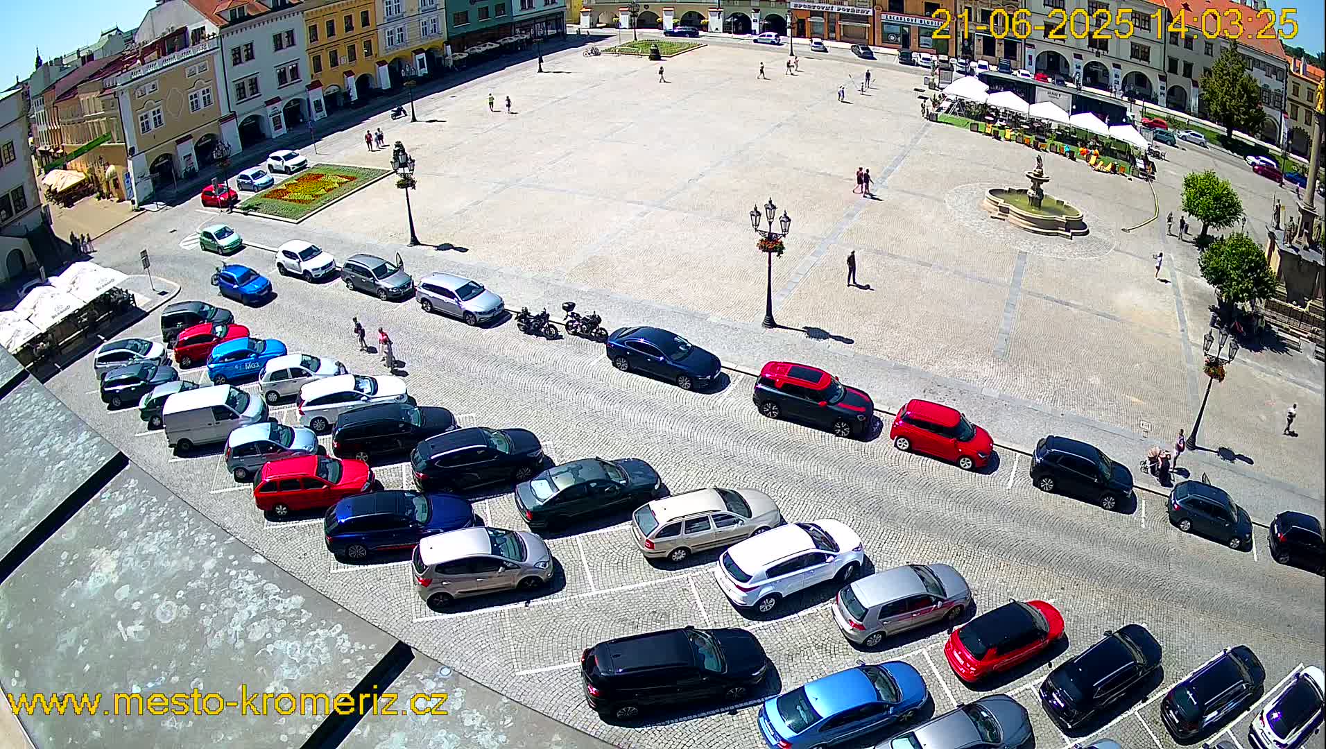 A town square with a fountain and several parked cars is seen from above on a sunny day.