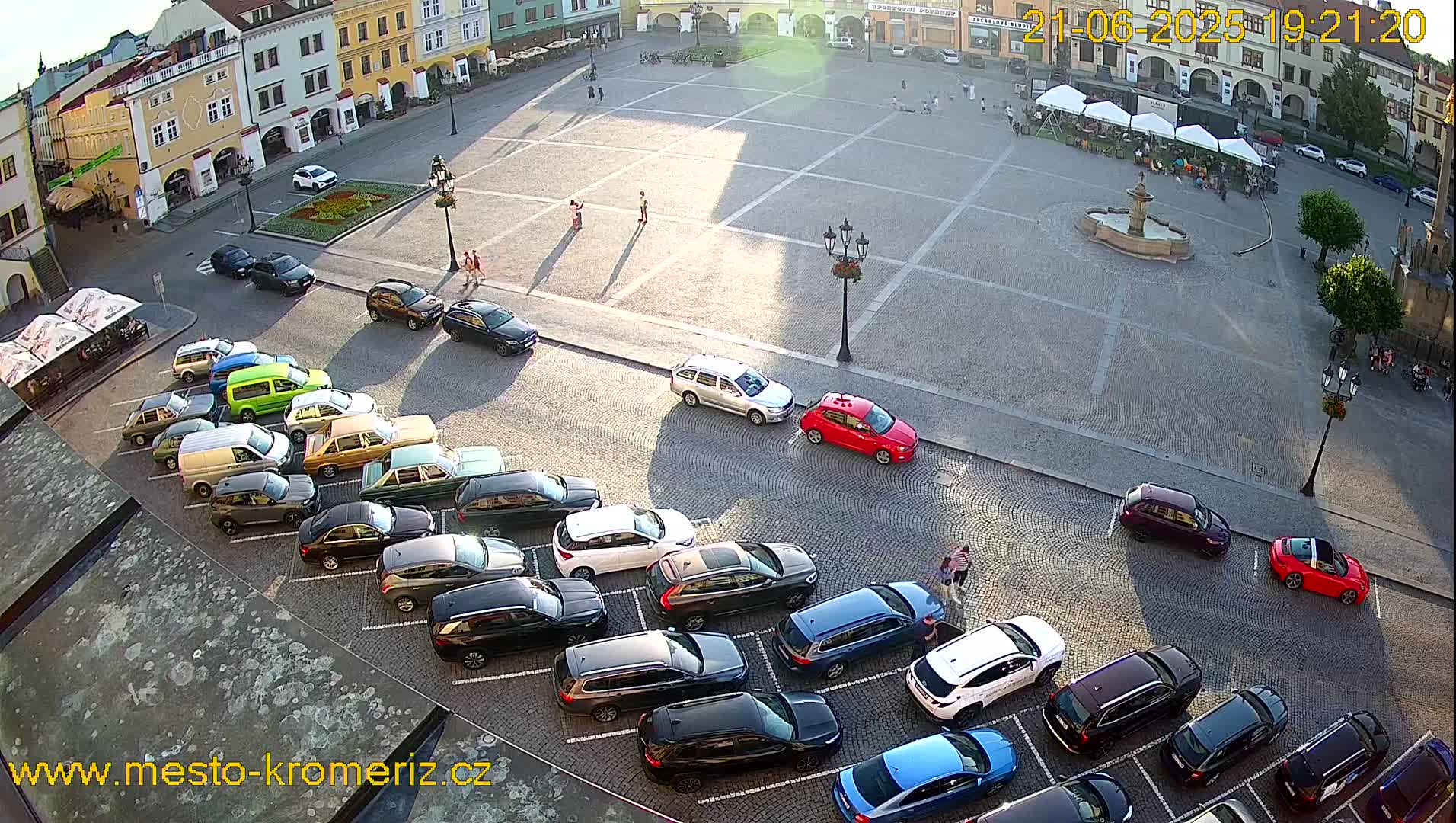 A sunny town square with a fountain, surrounded by parked cars and buildings, with several people walking around.