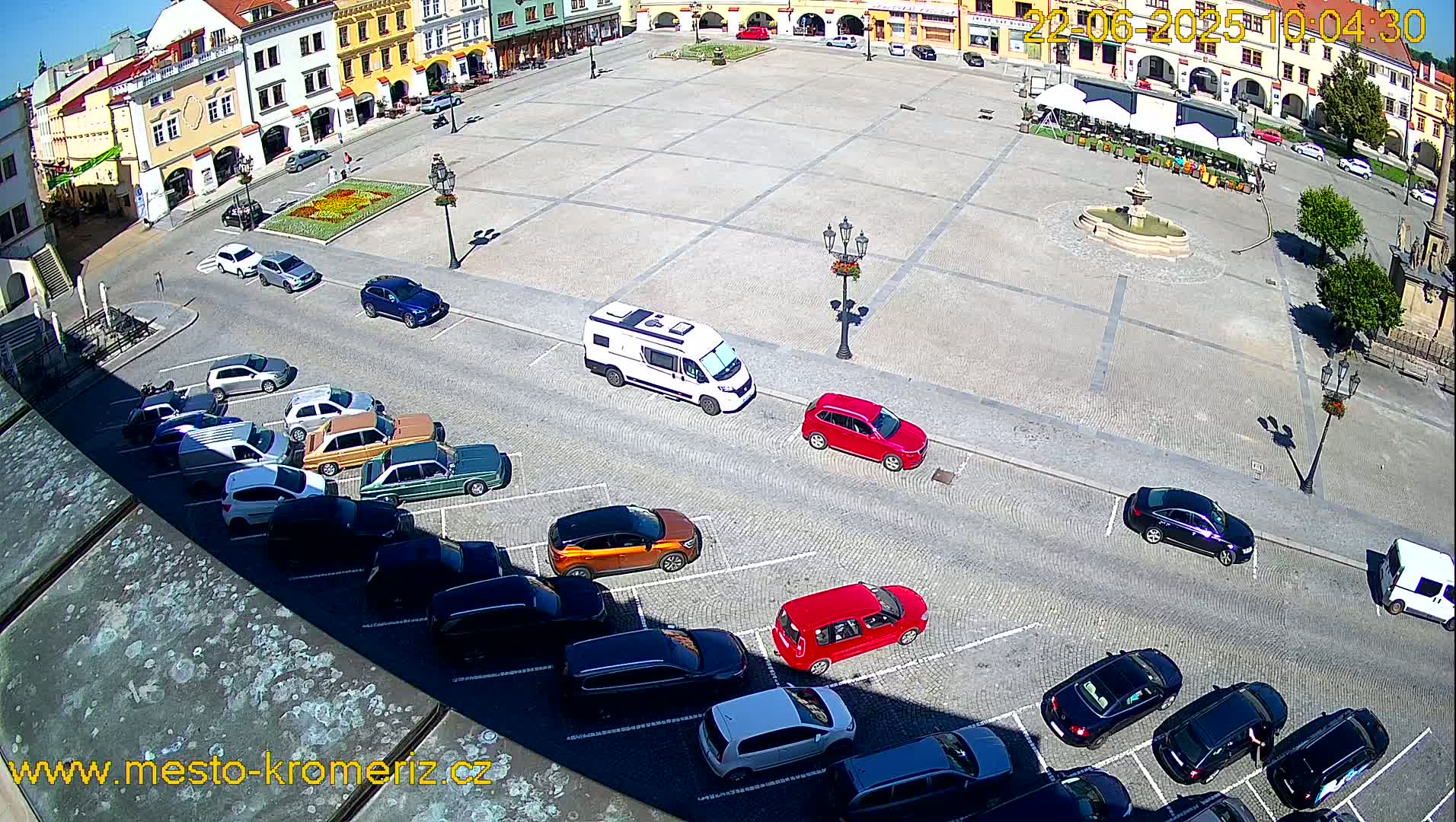 A large town square, under sunny conditions, is surrounded by buildings and has several cars parked along its edges.