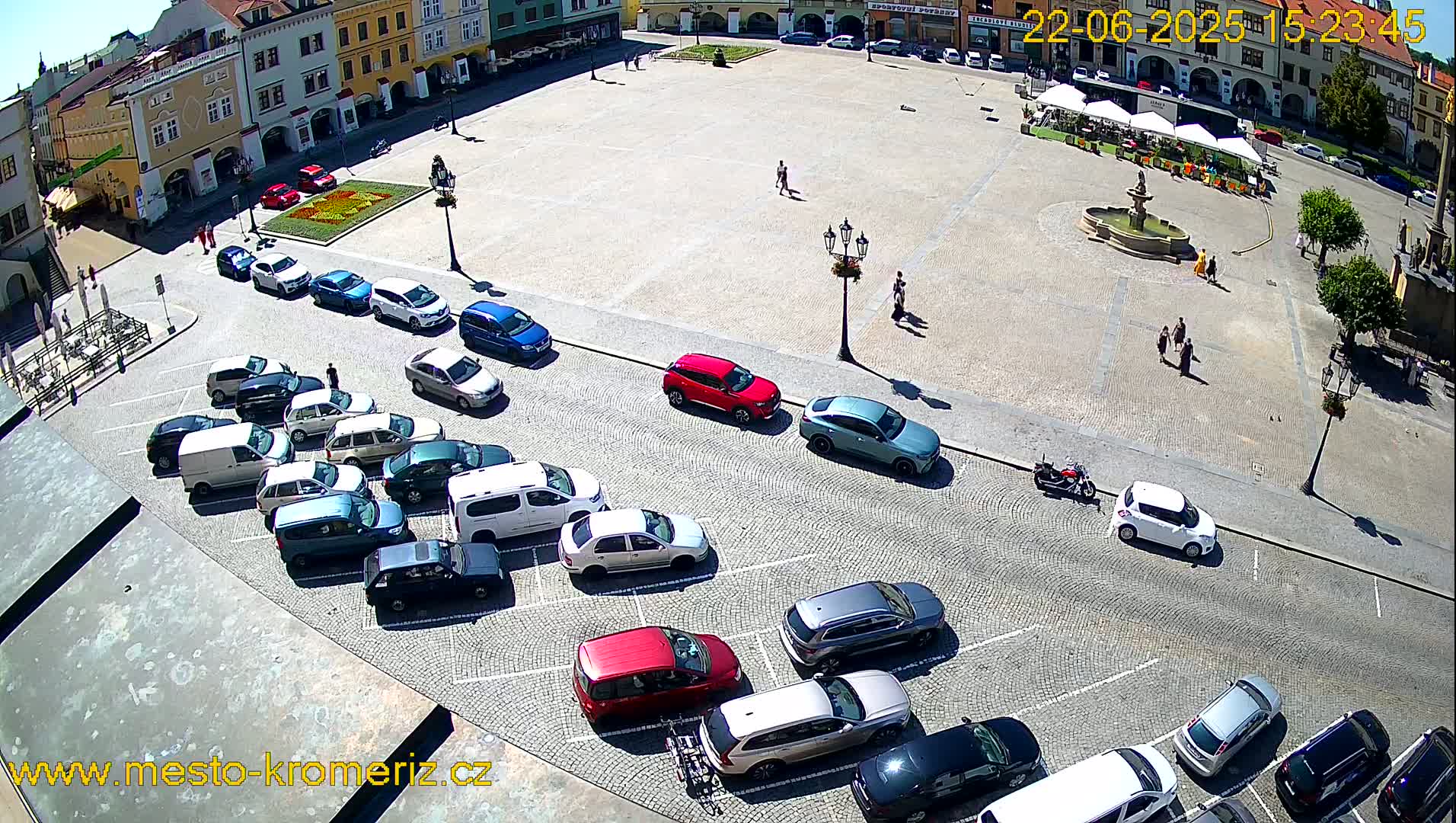A large town square, paved with cobblestones, is filled with parked cars and a few pedestrians on a sunny day.