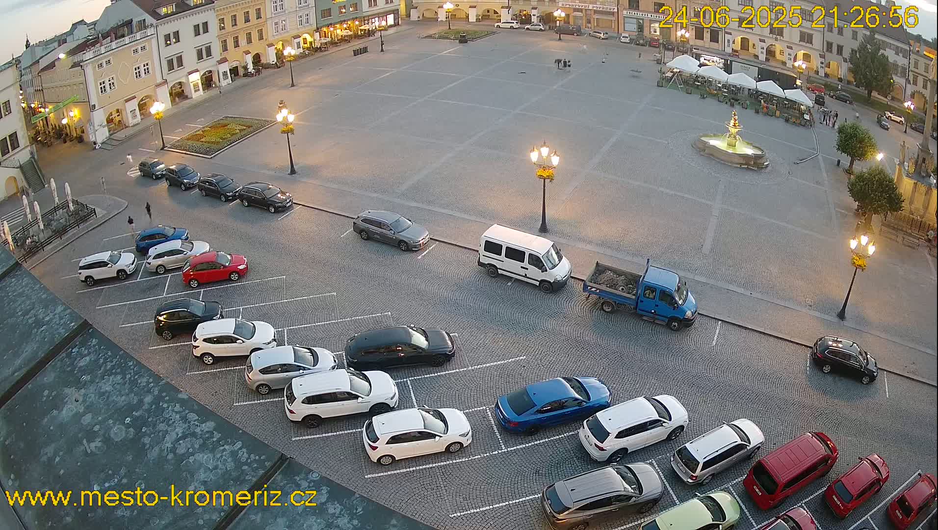 A town square at dusk with a fountain in the center, surrounded by parked cars and buildings, under a clear sky.