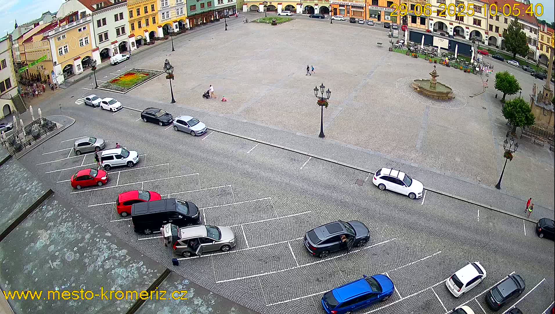 A cobblestone town square with a fountain and surrounding buildings, featuring a parking lot with several cars parked, under a partly cloudy sky.