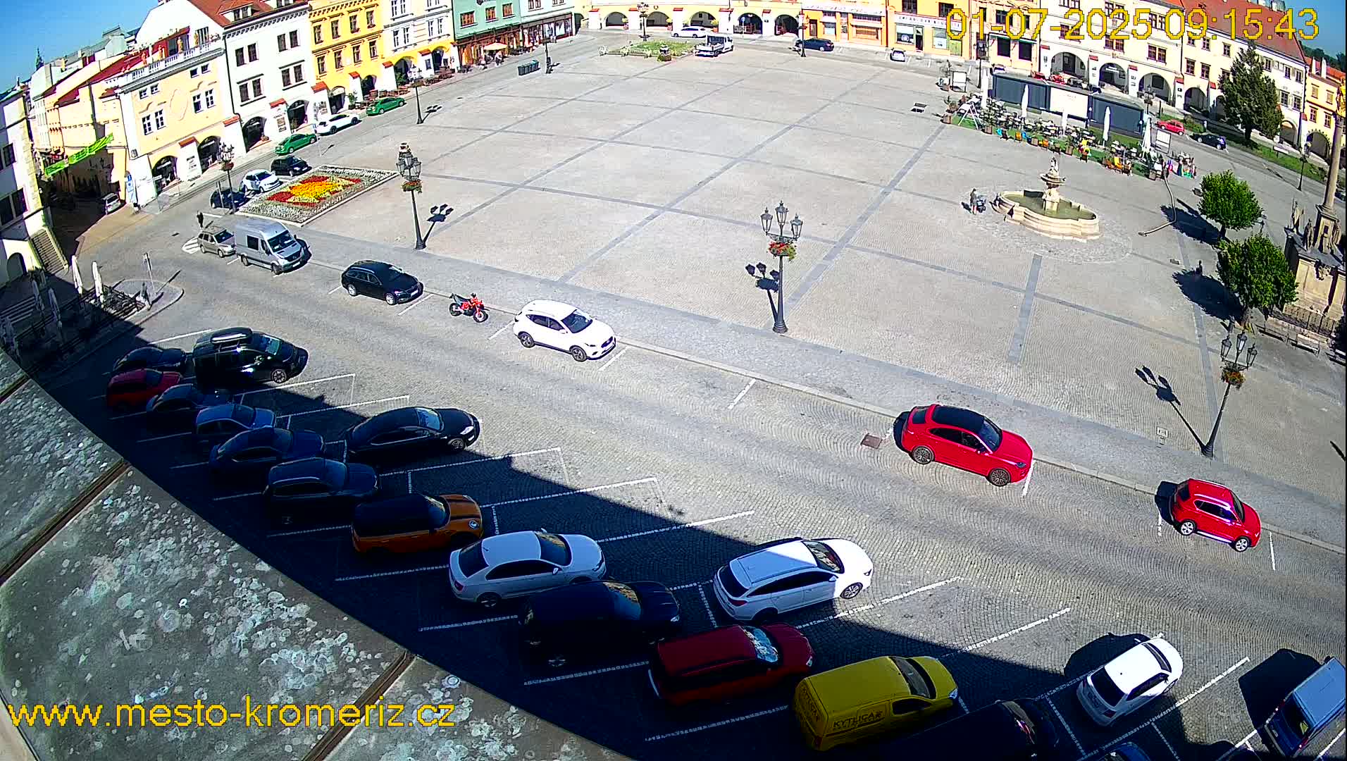A town square with a fountain is surrounded by parked cars and buildings under a sunny sky.