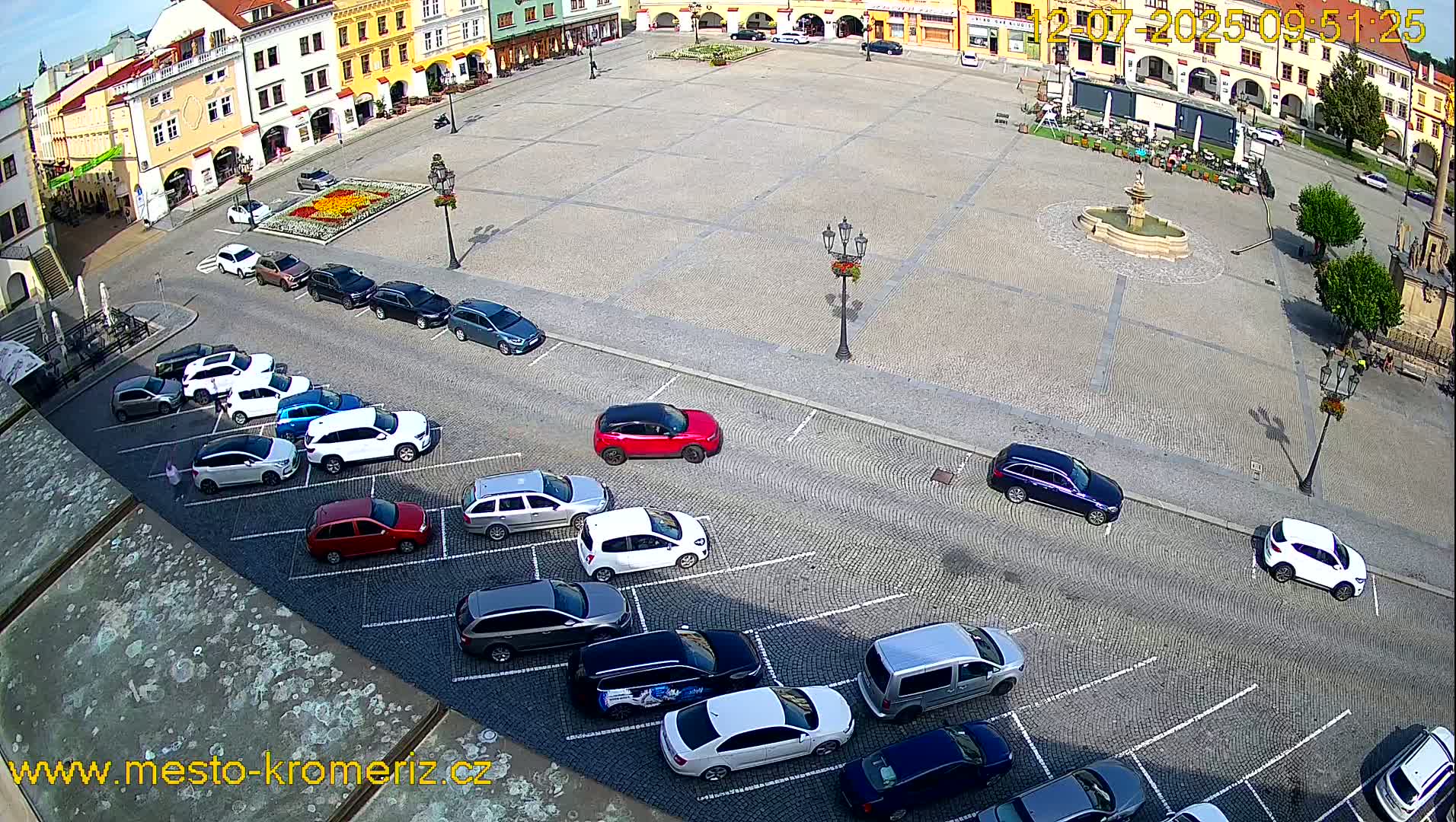 A large town square with a fountain in the center, surrounded by buildings and cars parked along the edges on a sunny day.